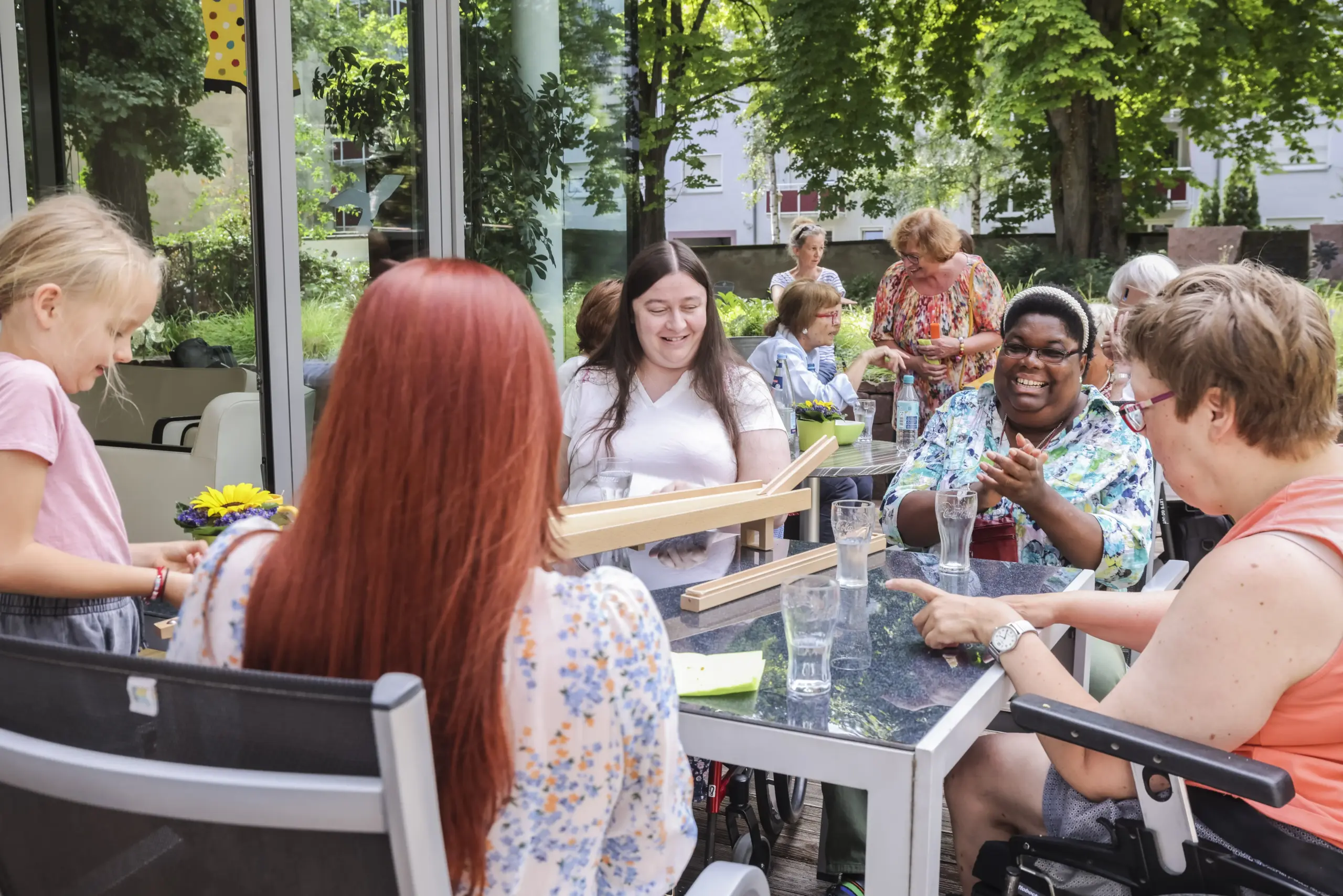 Vier Frauen sitzen rund um einen Tisch im Garten des Friedensheims. Auf dem Tisch steht eine Murmelbahn mit der ein Mädchen spielt. Im Hintergrund sitzen weitere Personen an Tischen.