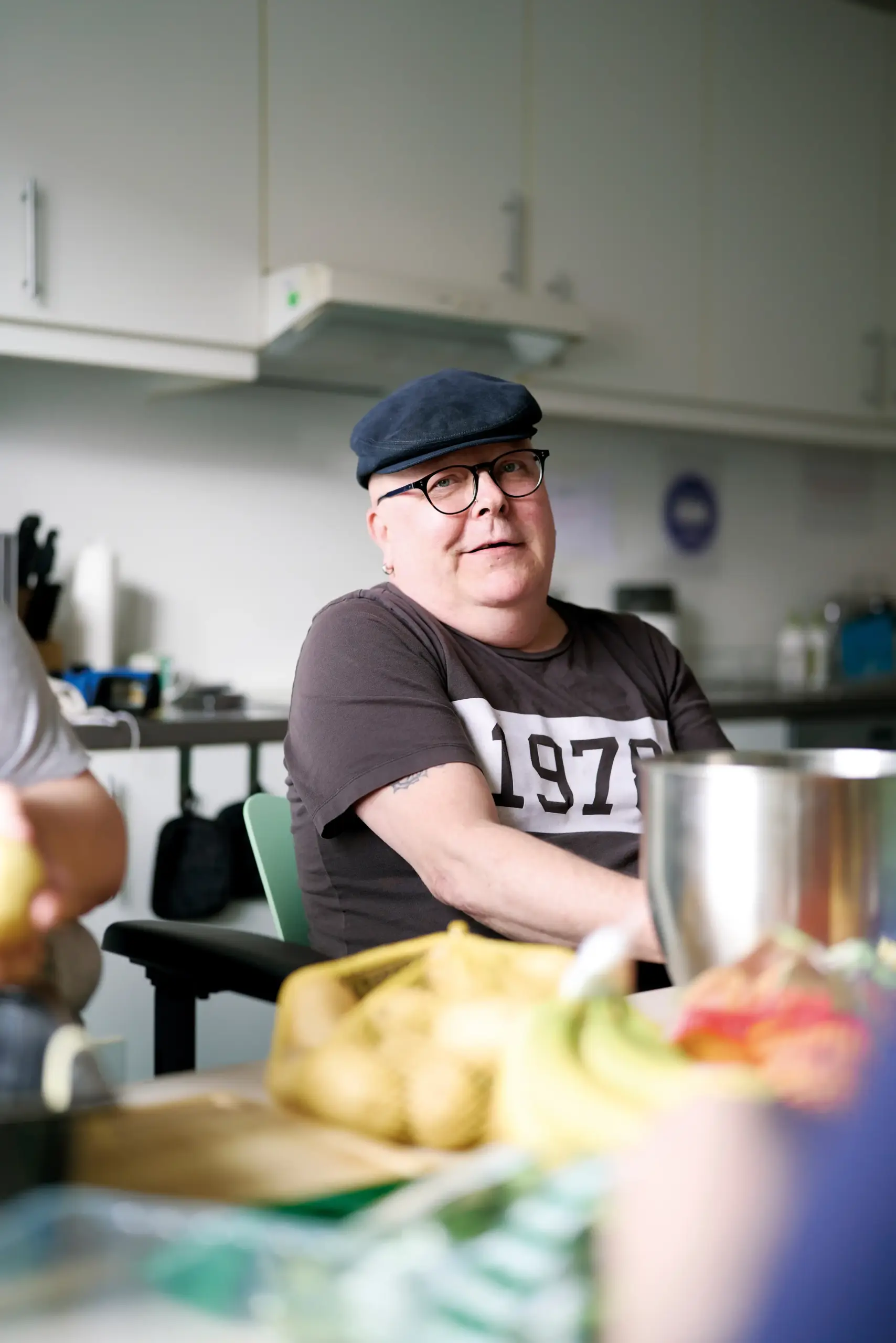 Ein Mann sitzt an einem Tisch. Er hat eine schwarze Schiebermütze auf, trägt eine Brille und ein braunes T-Shirt. Vor ihm auf dem Tisch liegt diverses Gemüse. Hinter ihm kann man vage eine Küche erkennen.