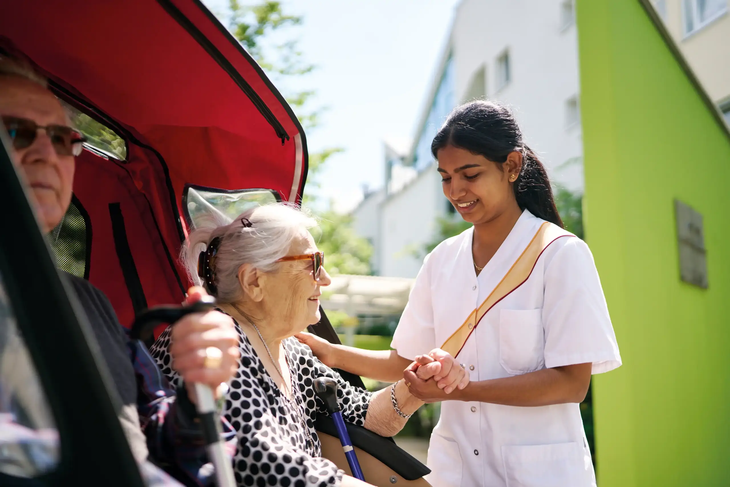 Eine junge Frau in Pflegerkleidung hilft einer älteren Frau aus einer Rikscha. Daneben sitzt ein älterer Mann. Im Hintergrund sieht man unscharf die Umrisse des Altenhilfezentrums Kalrsruhe-Nordost.