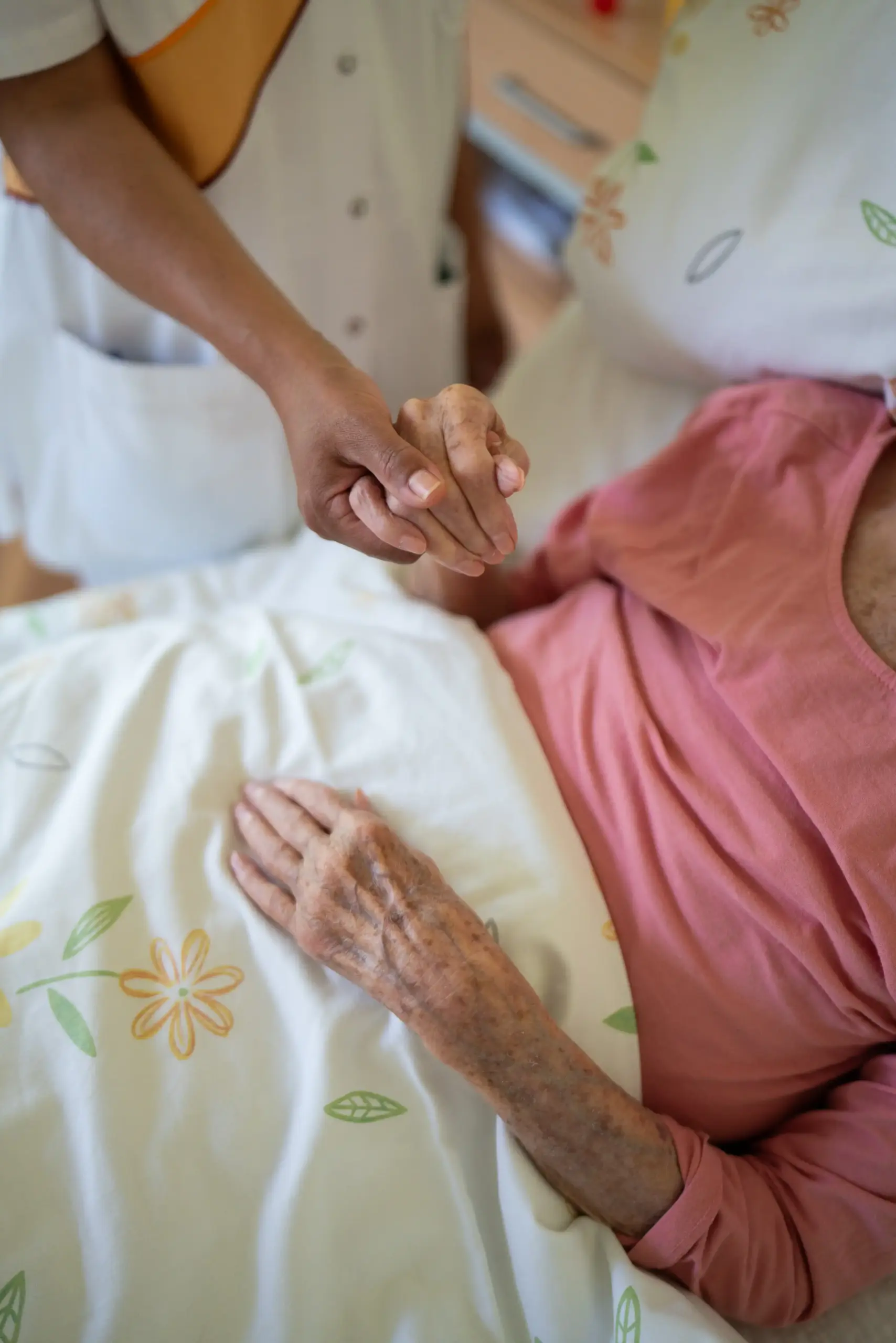 Quelle: BLV Eine junge Pflegerin hält die Hand einer älteren Frau, die im Bett liegt.