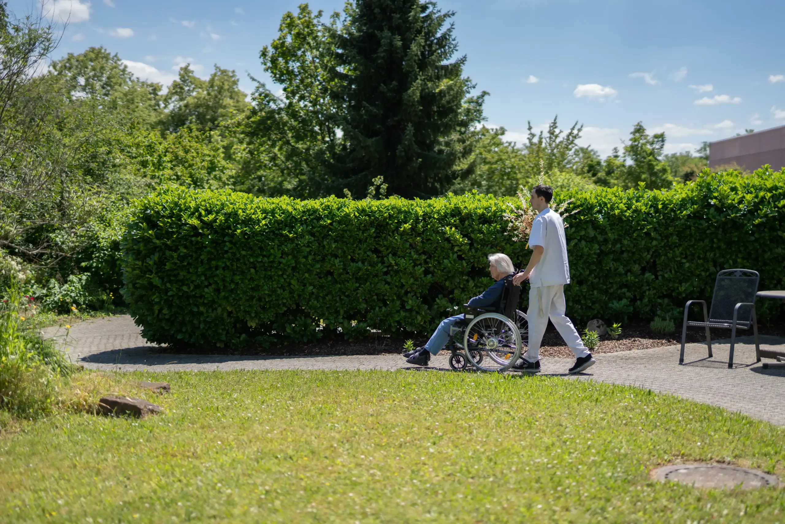 Quelle: BLV Eine junger Mann schiebt eine ältere Frau in einem Rollstuhl durch den Garten des Altenhilfezentrums Karlsruhe-Nordost.