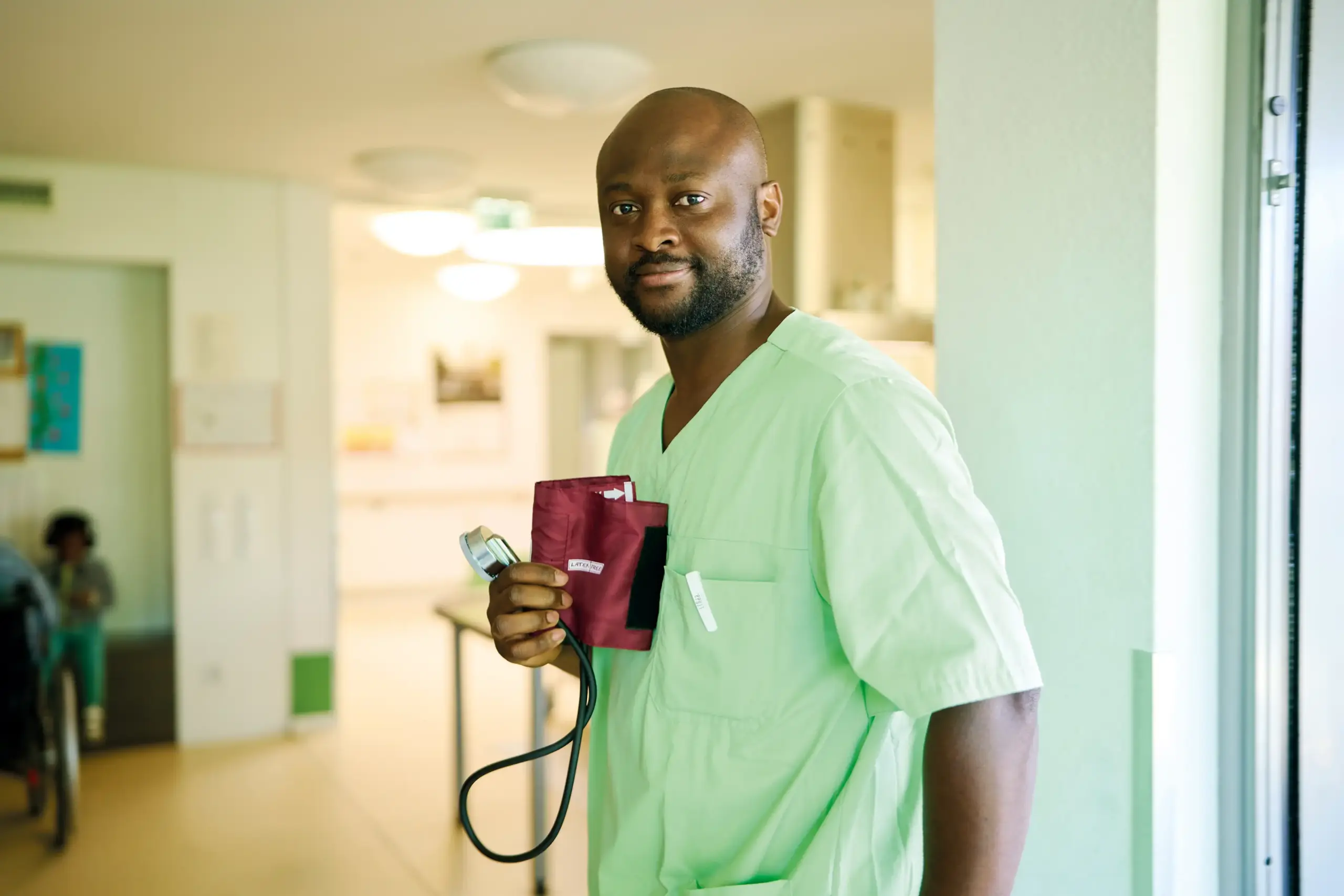 Ein Mann in grüner Pflege-Kleidung. In der Hand hält er ein Gerät, um den Blutdruck zu messen.