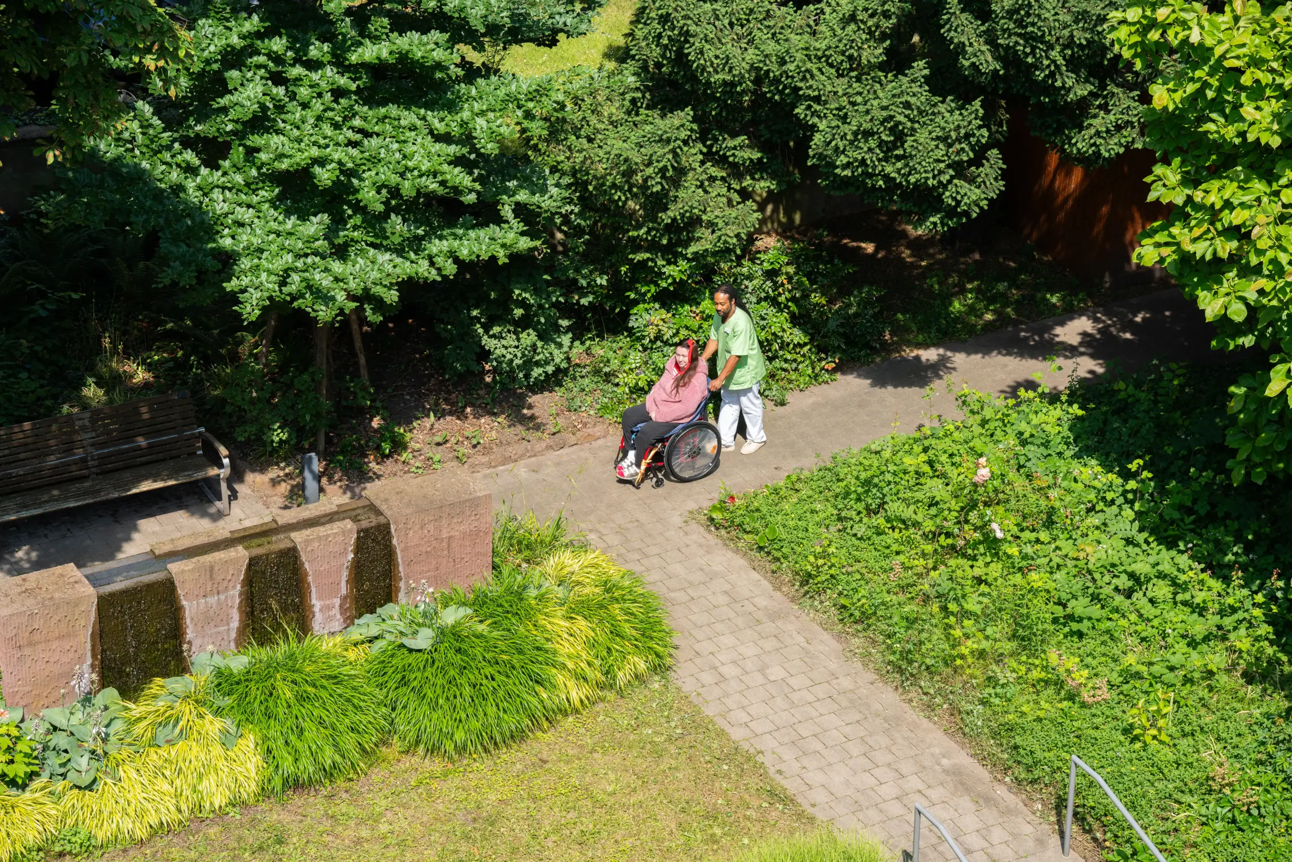 Quelle: BLV Ein Mann in grüner Pflegekleidung schiebt eine jüngere Frau in einem Rollstuhl im Garten des Friedensheims.