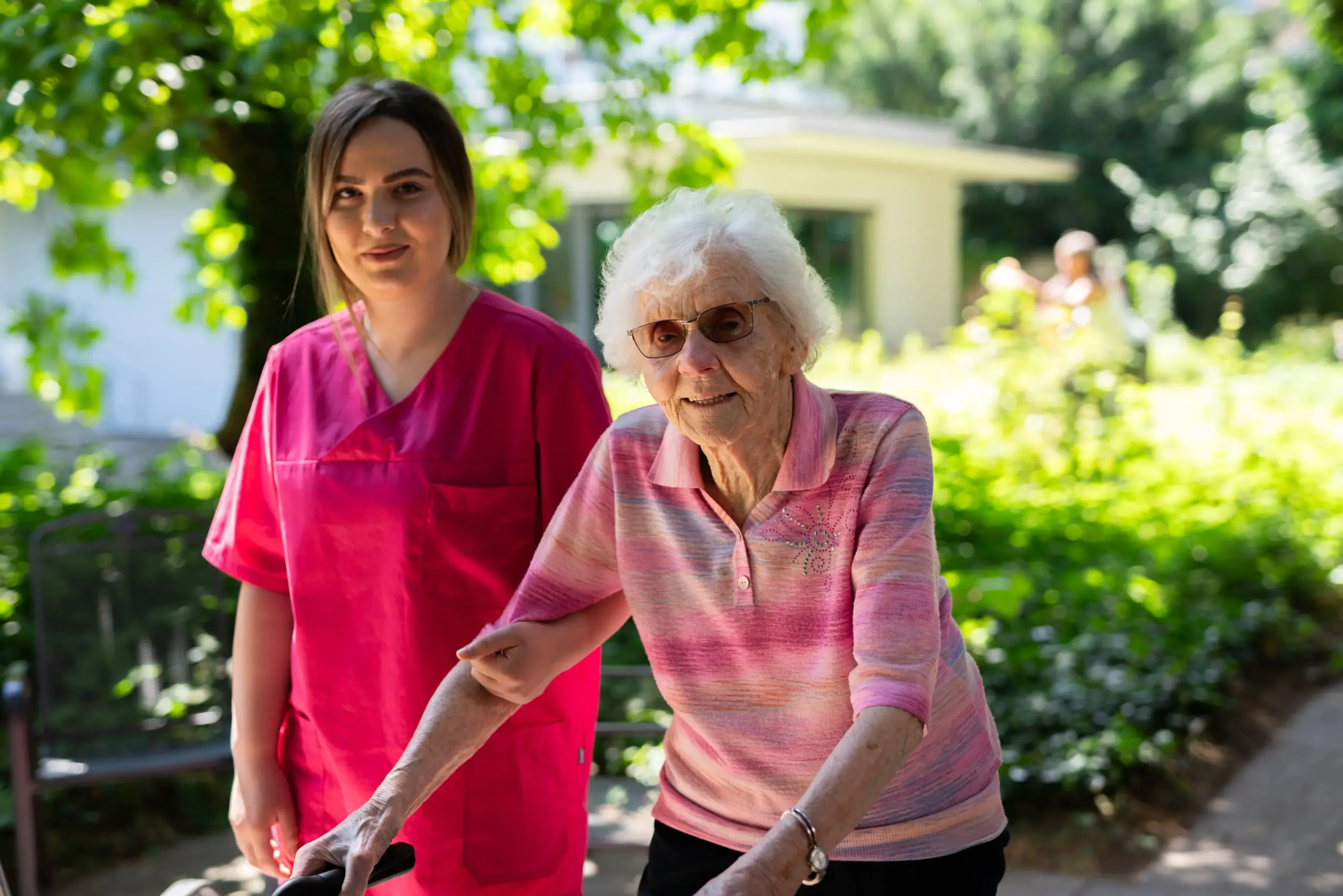 Quelle: BLV Eine jüngere Frau und eine ältere Frau stehen nebeneinander im Garten des Friedensheims. Die ältere Frau hat ihre Hände an einem Rollator, die jüngere Frau trägt rote Pflegekleidung und hat sich bei ihr untergehakt. Sie lächeln in die Kamera.