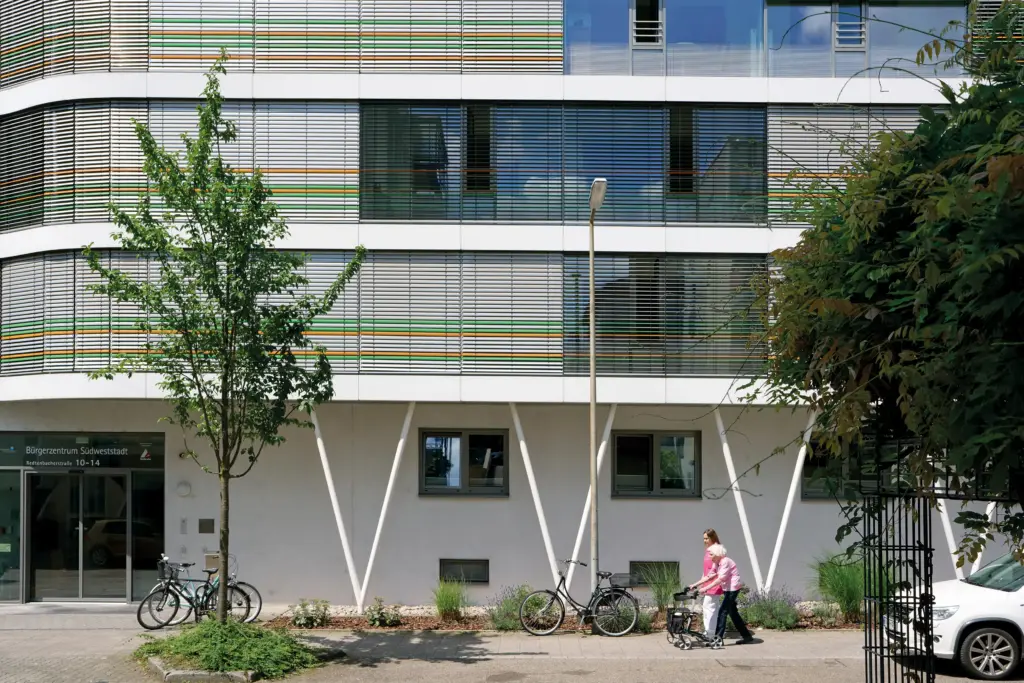 Das Friedensheim von der Straße aus betrachtet. Eine Frau mit Rollator und eine Pflegerin laufen davor über den Gehweg.