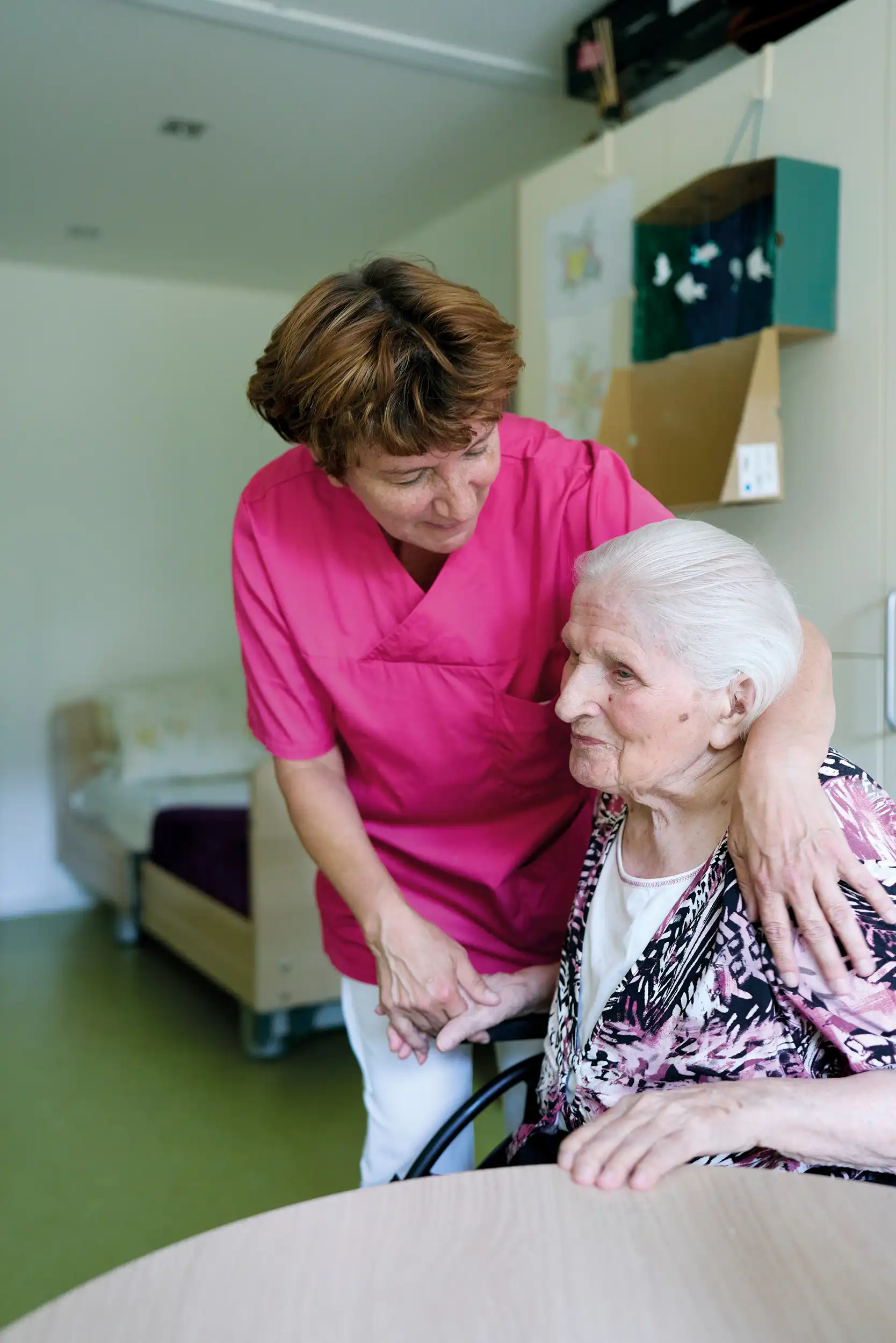 Quelle: BLV Eine ältere Frau sitzt an einem Tisch. Eine andere Frau in roter Pflegekleidung steht neben ihr und legt den Arm um sie. Im Hintergrund sieht man ein Krankenbett.