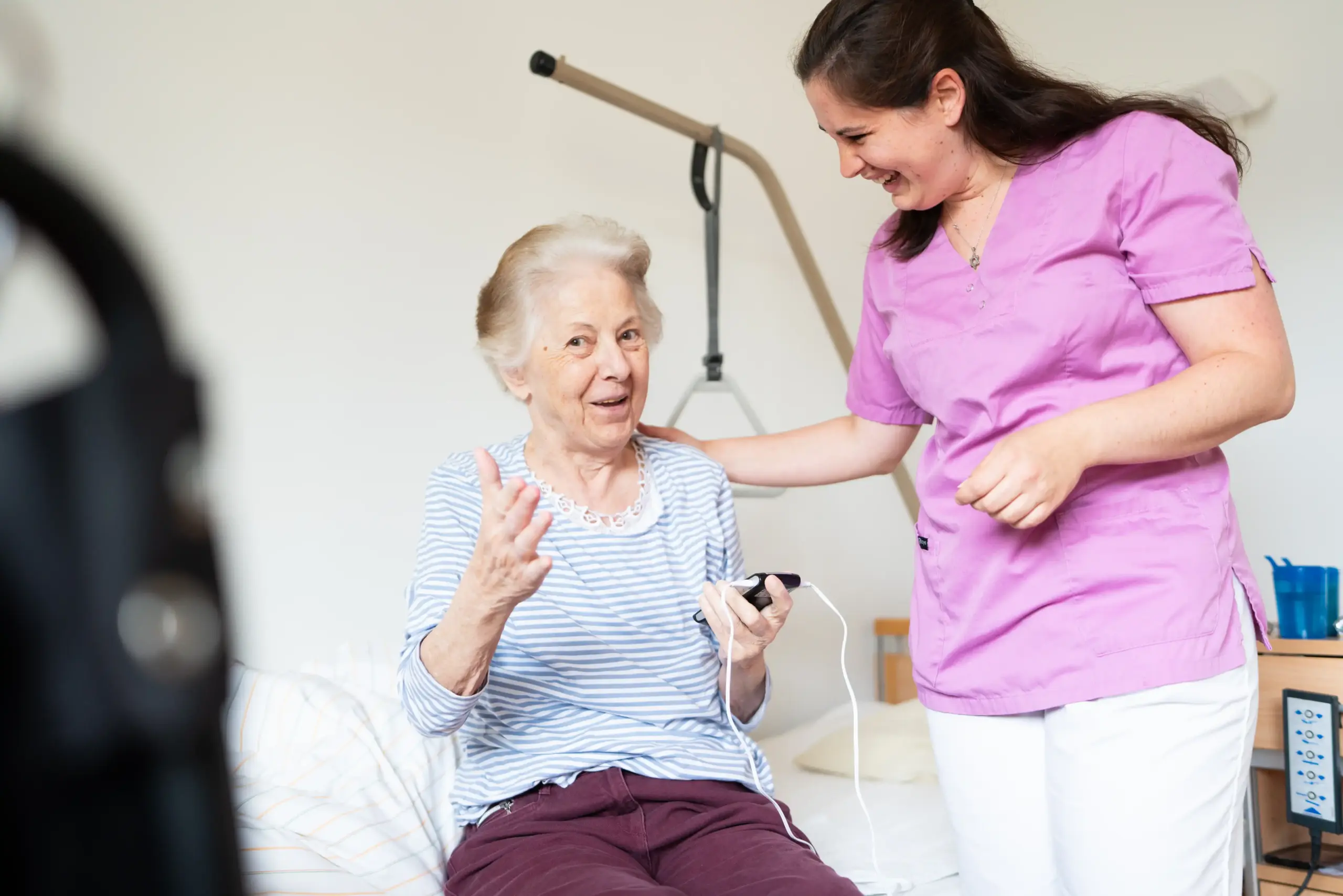 Eine ältere Frau sitzt auf einem Bett. In ihren Händen hält sie ein Gerät, in dem zwei Kabel stecken. Neben ihr steht eine Pflegerin in violetter Pflegekleidung. Sie hat ihre Hand auf die Schulter der älteren Frau gelegt. Beide lachen.