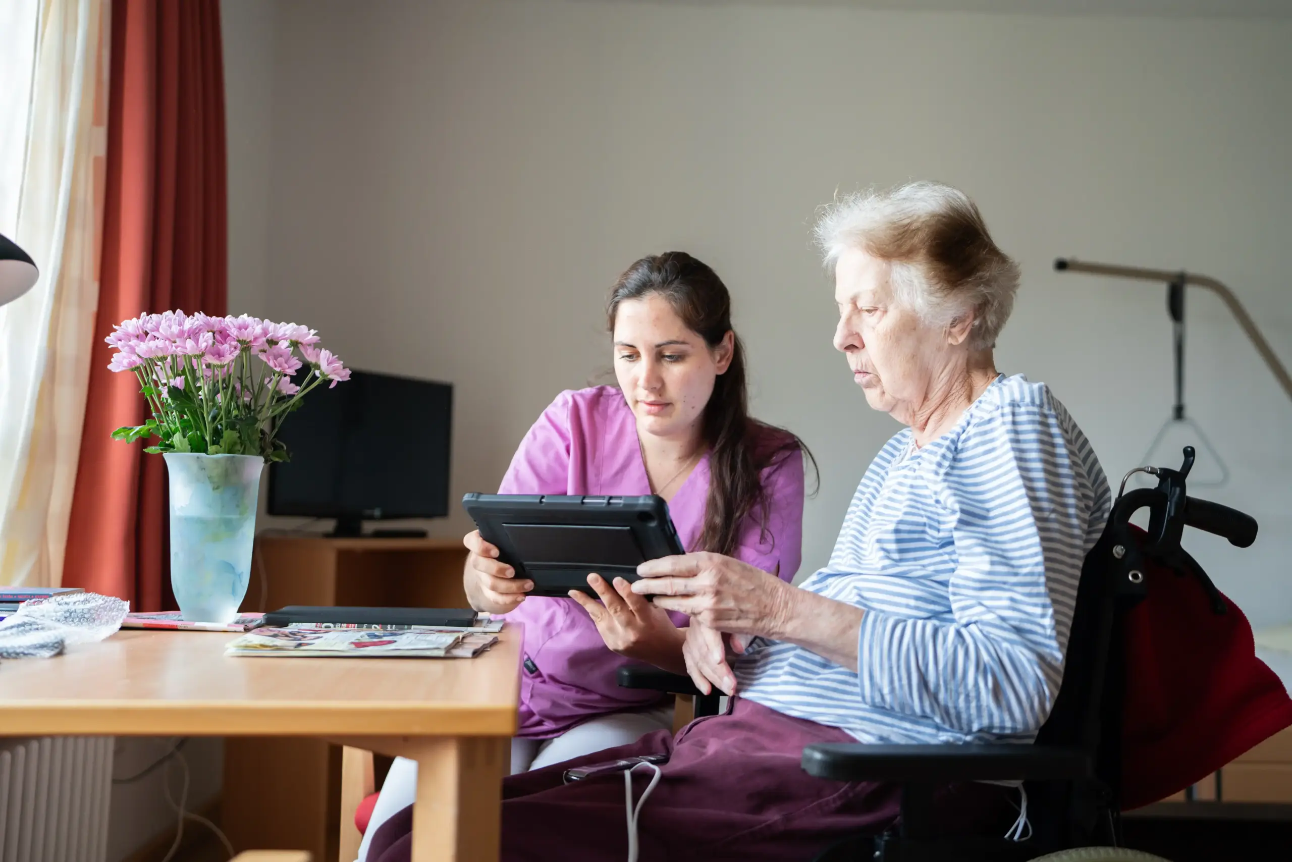 Quelle: BLV Eine ältere Frau und eine jüngere Frau schauen gemeinsam auf ein Tablet. Die ältere Frau sitzt in einem Rollstuhl. Die jüngere Frau trägt violette Pflegekleidung. Im Hintergrund stehen ein Fernseher und ein Bett.