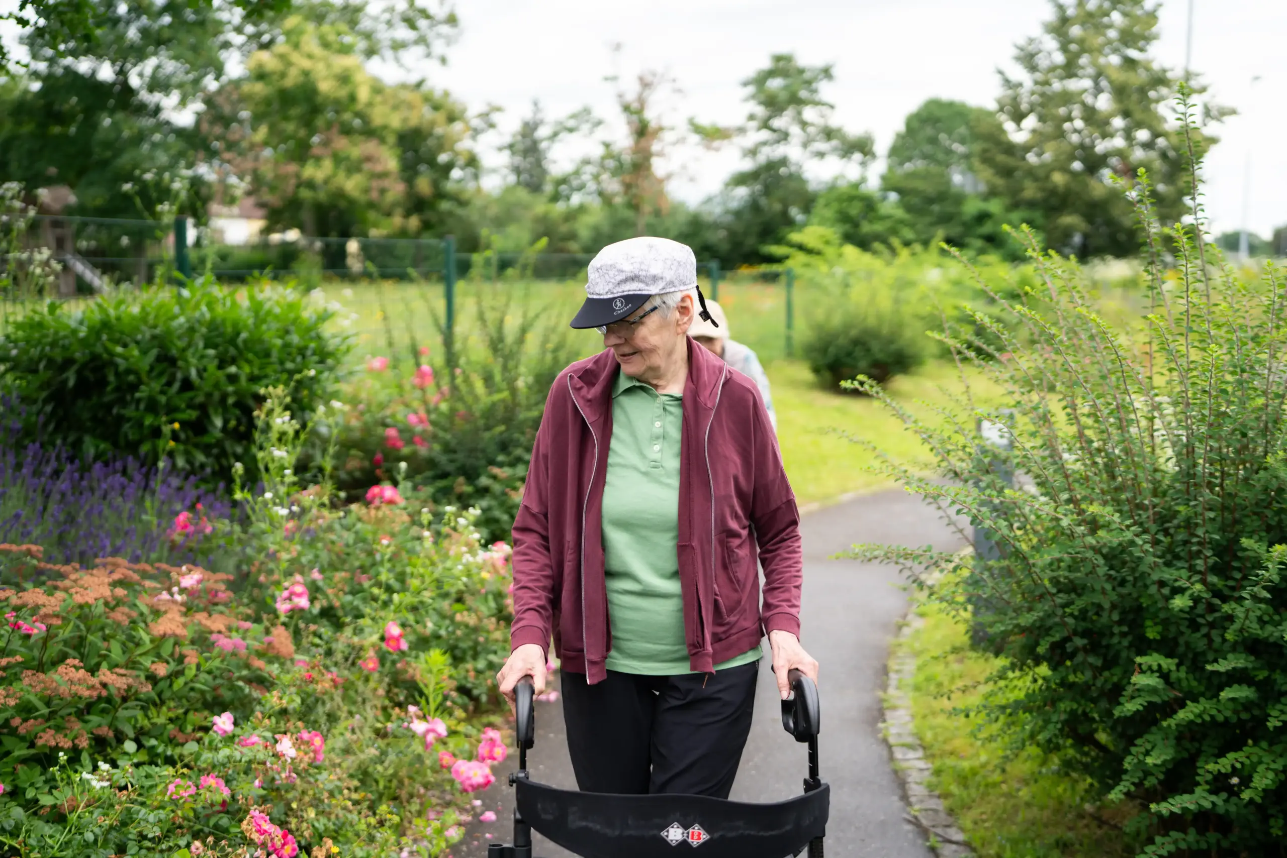 Zwei Frau spazieren hintereinander mit ihren Rollatoren durch den Garten des Hauses Karlsruher Weg. Sie betrachten die Blumen neben dem Weg.