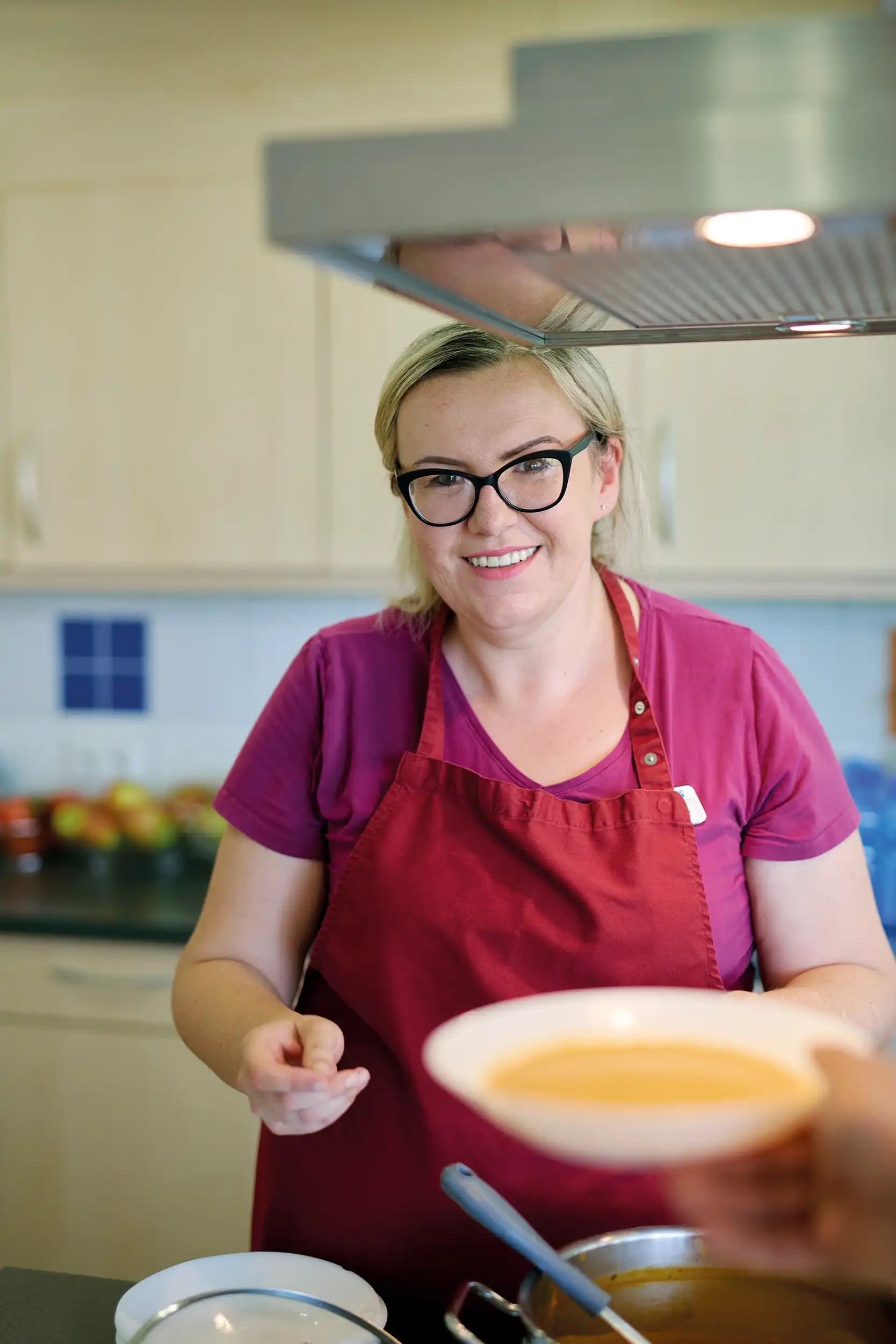Eine Frau reicht einen Teller Suppe. Sie trägt ein violettes Shirt und eine rote Schürze. Vor Ihr auf dem Herd steht ein Topf mit Suppe.