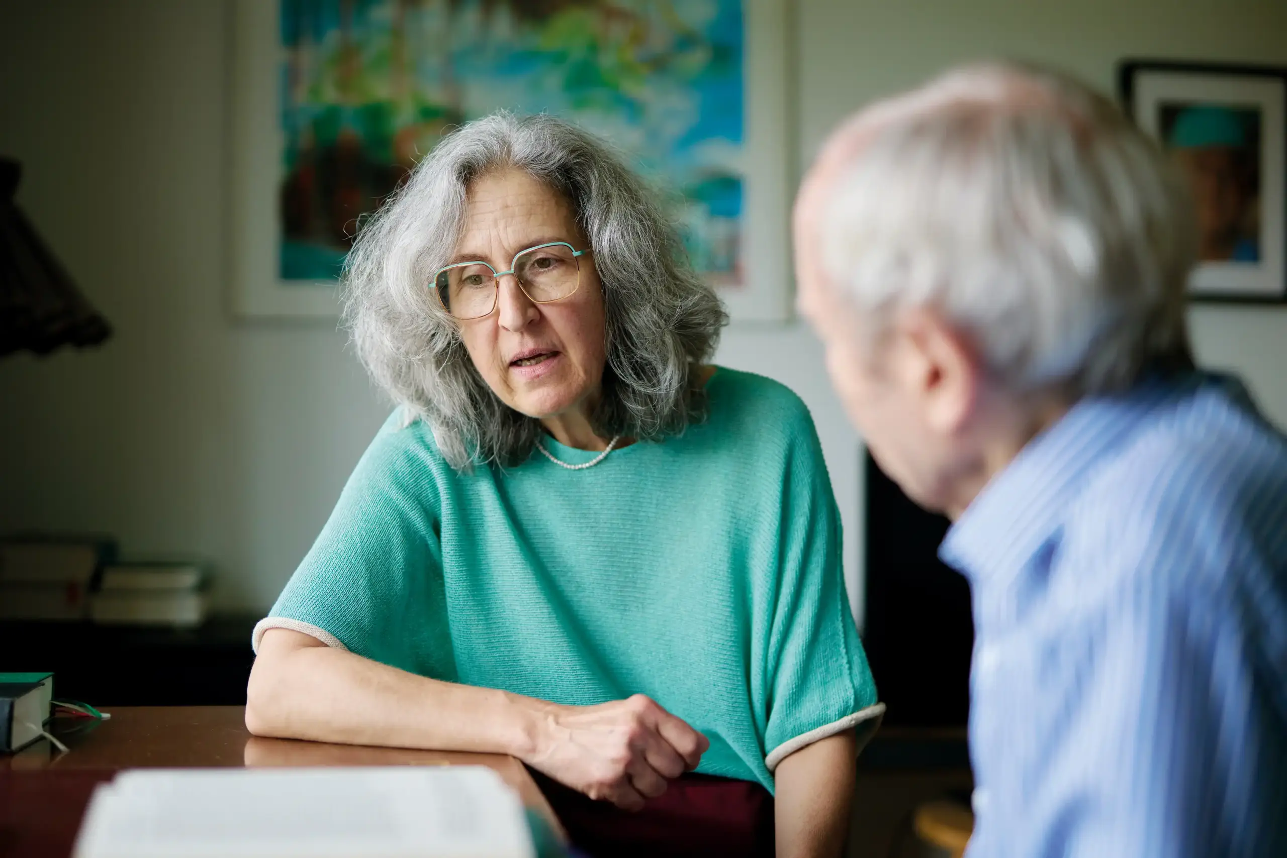Eine Frau sitzt an einem Tisch. Sie trägt einen türkis-farbenen Pullover. Die Frau spricht mit einem älteren Mann.