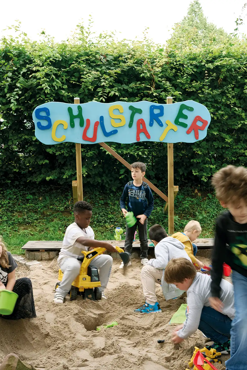 Sieben Kinder spielen in einem Sandkasten. Dahinter steht ein Schild auf dem in großen, bunten Buchstaben 