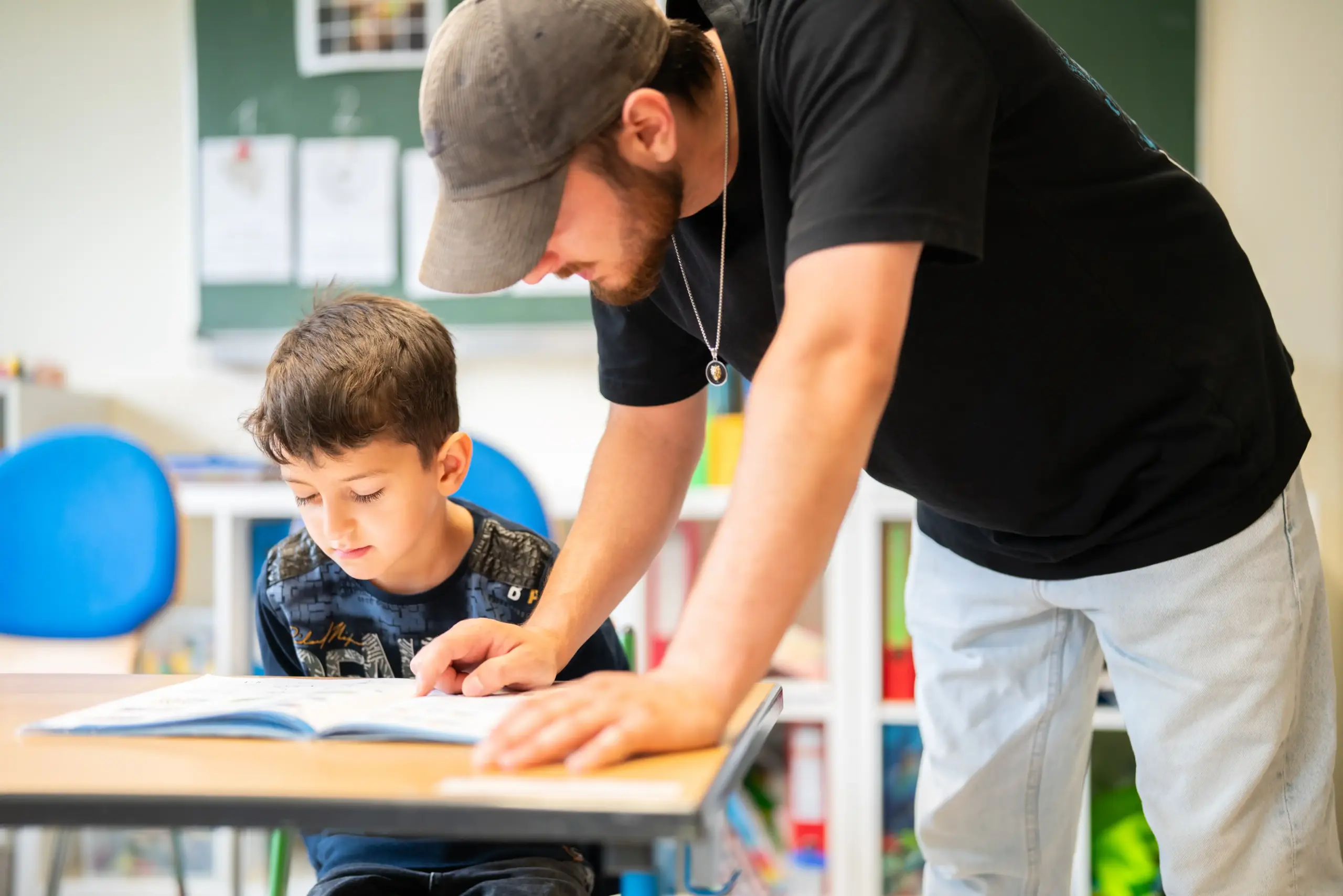 Ein Junge sitzt an einem Schultisch und liest in seinem Heft. Ein junger Mann steht neben ihm und zeigt ihm etwas darin. Im Hintergrund sind eine Tafel und verschiedene Ordner zu erkennen.