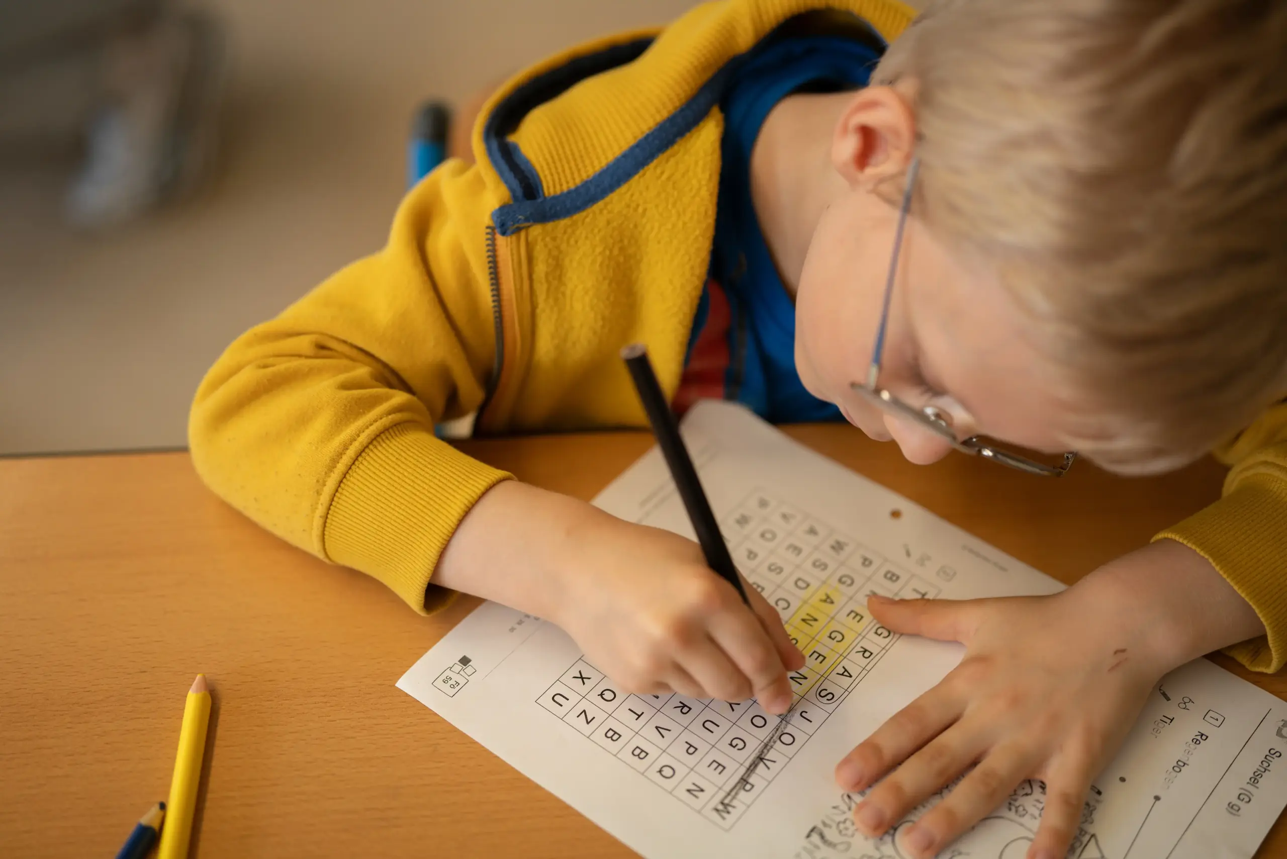 Ein Junge sitzt an einem Tisch und löst seine Hausaufgaben. Er trägt eine Brille und eine gelbe Jacke.