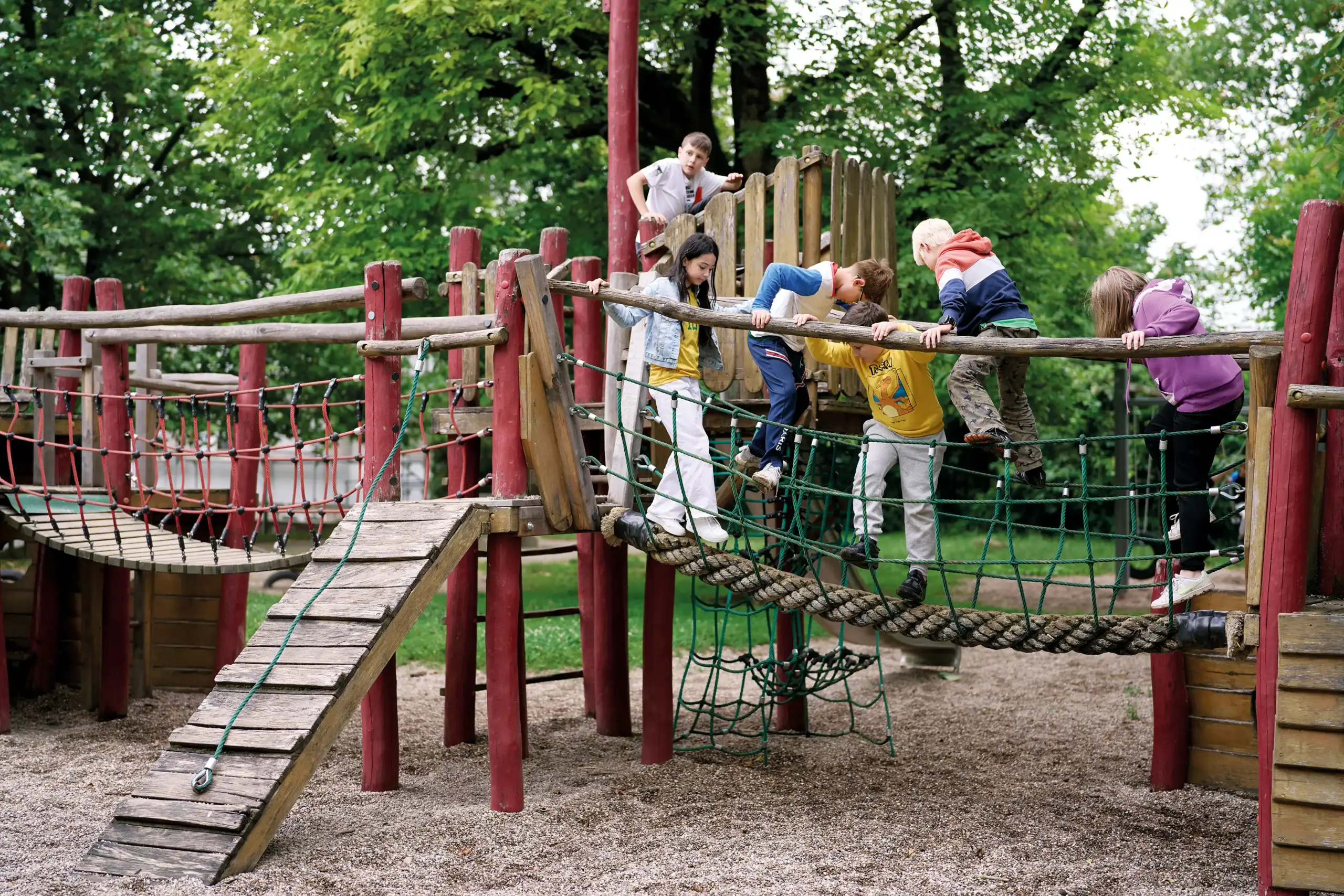 Sechs Kinder spielen auf dem Klettergerüst eines Spielplatzes. Fünf von ihnen klettern auf einer Hängebrücke herum.