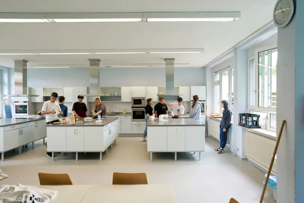 Neun Personen arbeiten in einer Großküche. Von den drei Kücheninseln wird an zwei Kücheninseln gearbeitet. An der einen mischt ein Junge mit einem Handrührgerät in einer Schüssel Teig an. An der anderen sind zwei Kinder damit beschäftigt, etwas in einer Pfanne anzubraten.