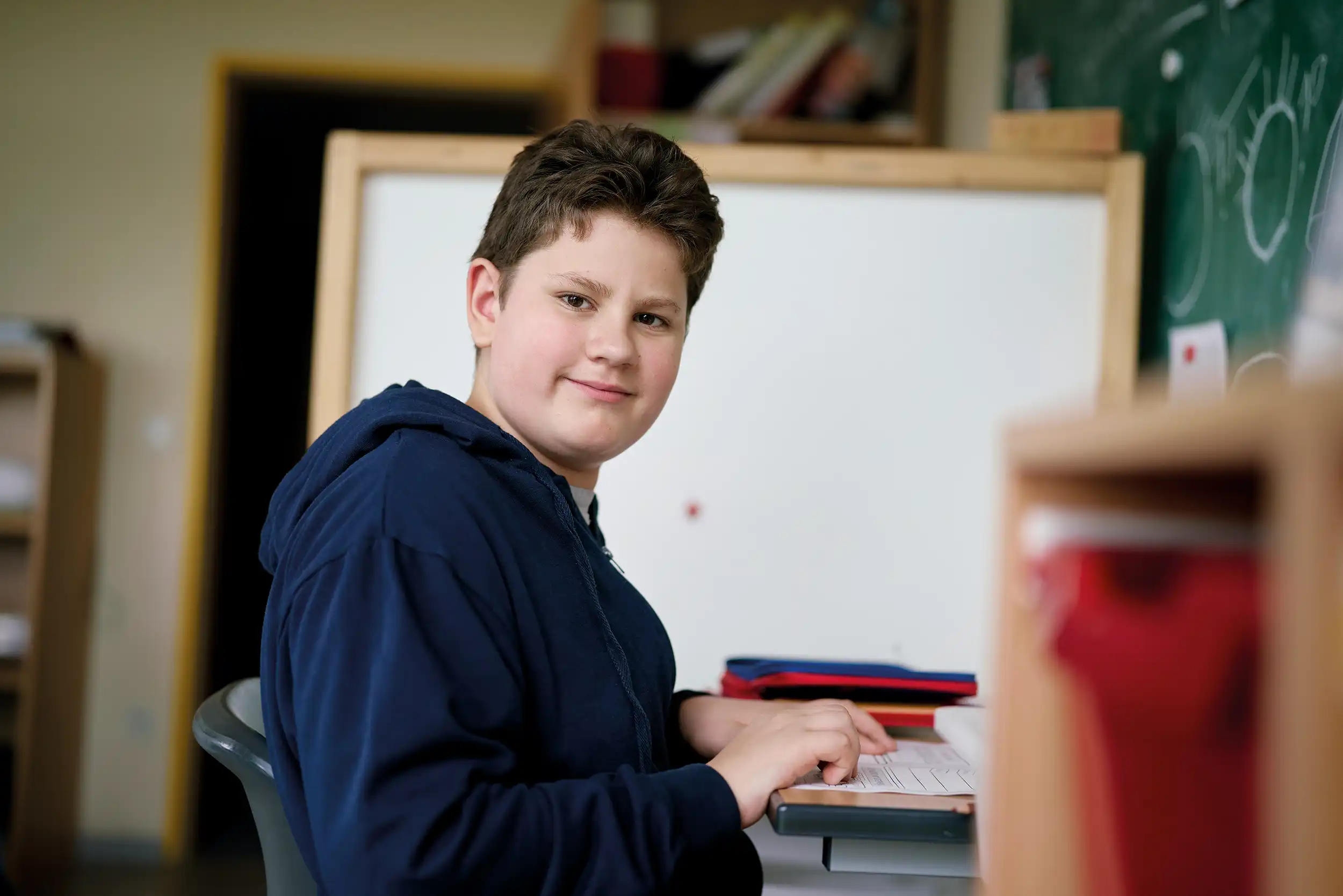 Ein Junge sitzt an einem Tisch. Vor ihm ein Blatt Papier. Er dreht sich zur Seite und lächelt in die Kamera.