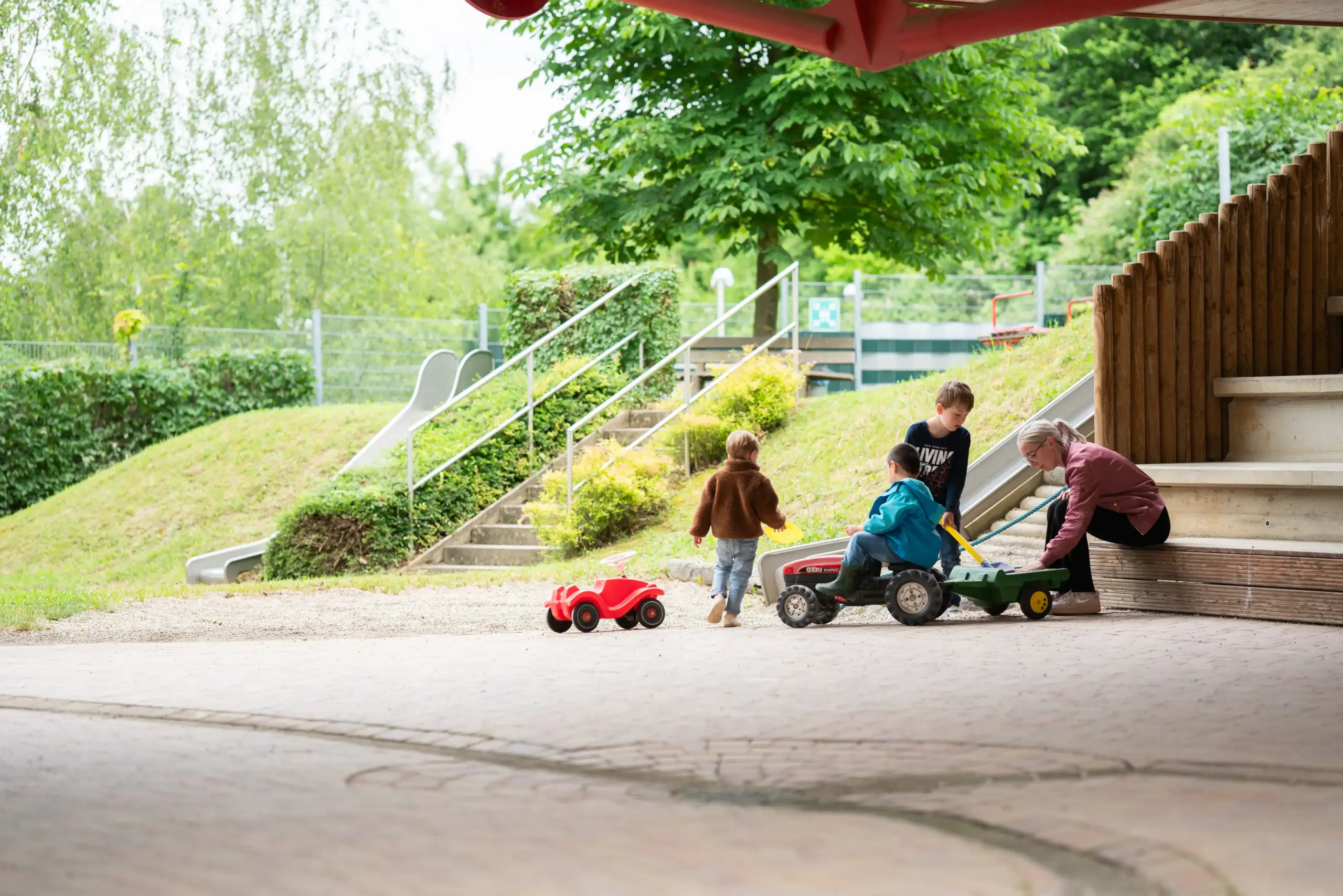 Drei Kinder spielen miteinander mit Spielfahrzeugen: einem Bobbycar und einem Traktor. Sie schaufeln Sand in den Anhänger des Traktors. Eine junge Frau sitzt daneben und hilft mit.