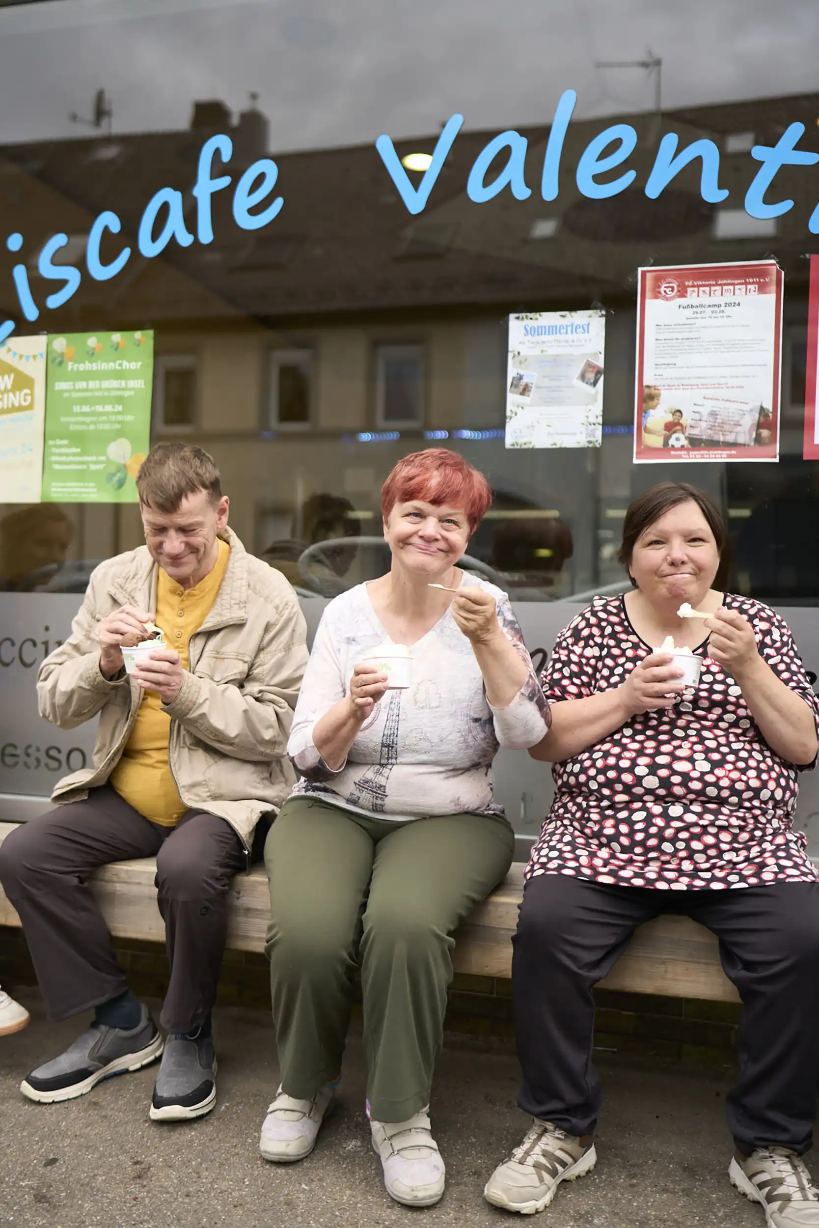 Drei Personen sitzen auf einer Bank und löffeln Eis aus kleinen Bechern.