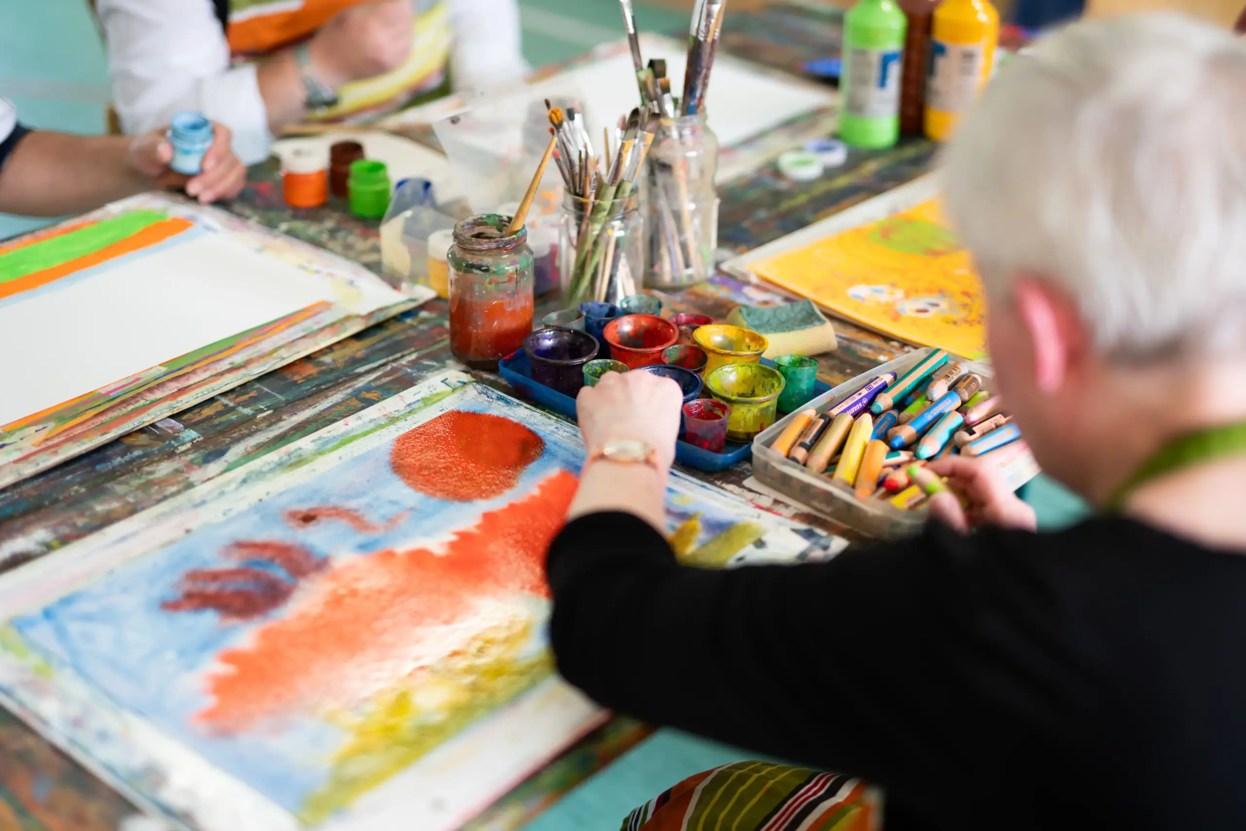 Eine Frau malt mit Wasserfarben. Auf dem Tisch vor ihr liegt das Papier, auf dem sie malt. Außerdem stehen auf dem Tisch Gläser mit Pinseln, kleine Farbbecher und Farbmalstifte.