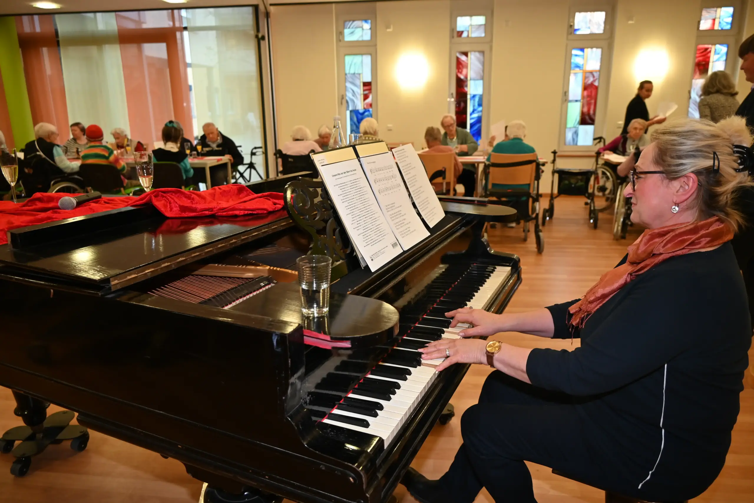 Quelle: BLV Eine Frau spielt auf einem Flügel Klavier. Im Hintergrund sitzen einige ältere Menschen an kleinen Tischen zusammen. Manche von ihnen haben Sekt auf dem Tisch stehen.
