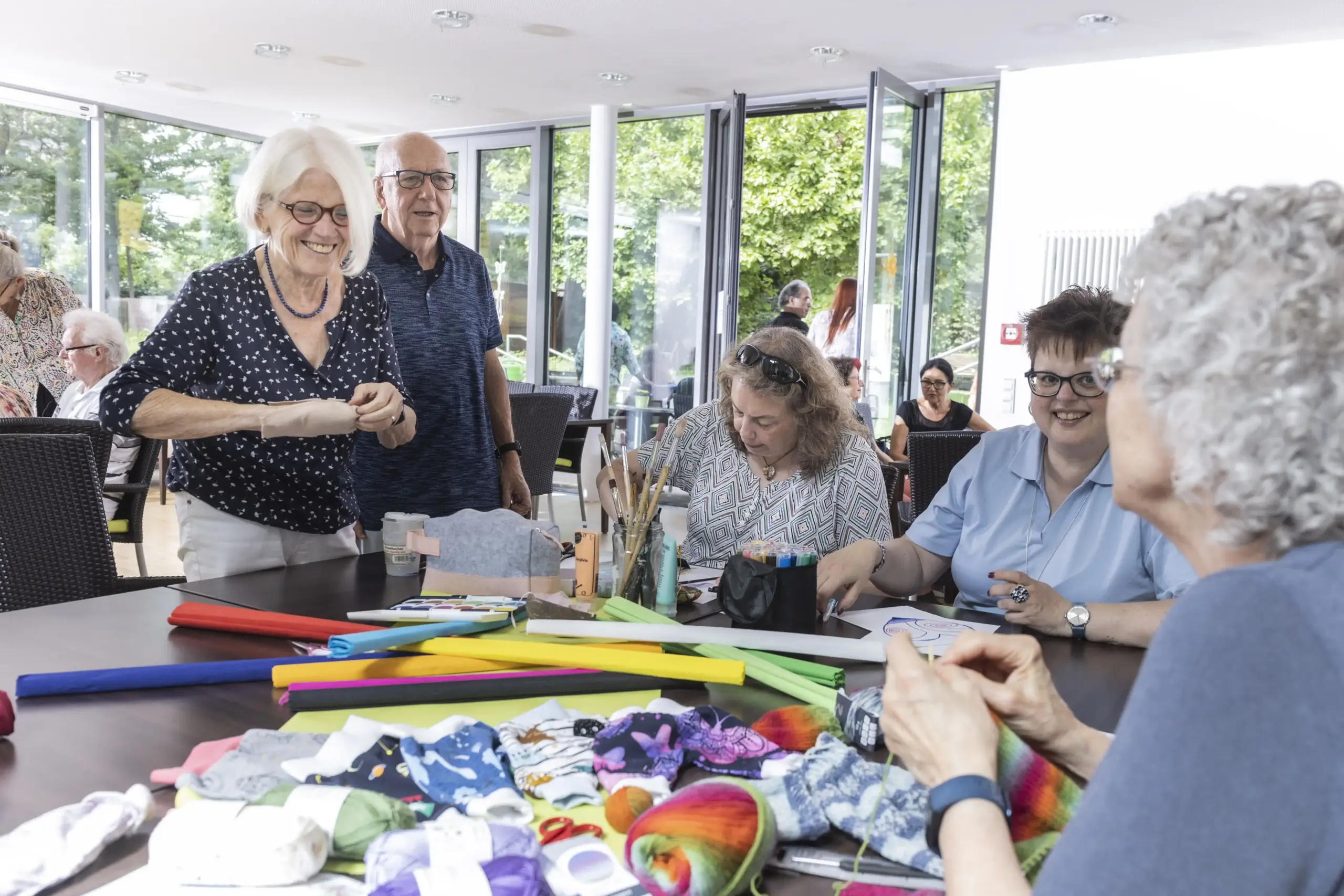 Quelle: BLV Vier Frauen sitzen rund um einen Tisch. Eine der Frauen strickt. Zwei andere malen. In der Mitte des Tisches liegt Krepp-Papier. Im Hintergrund sitzen weitere Menschen an Tischen und unterhalten sich.