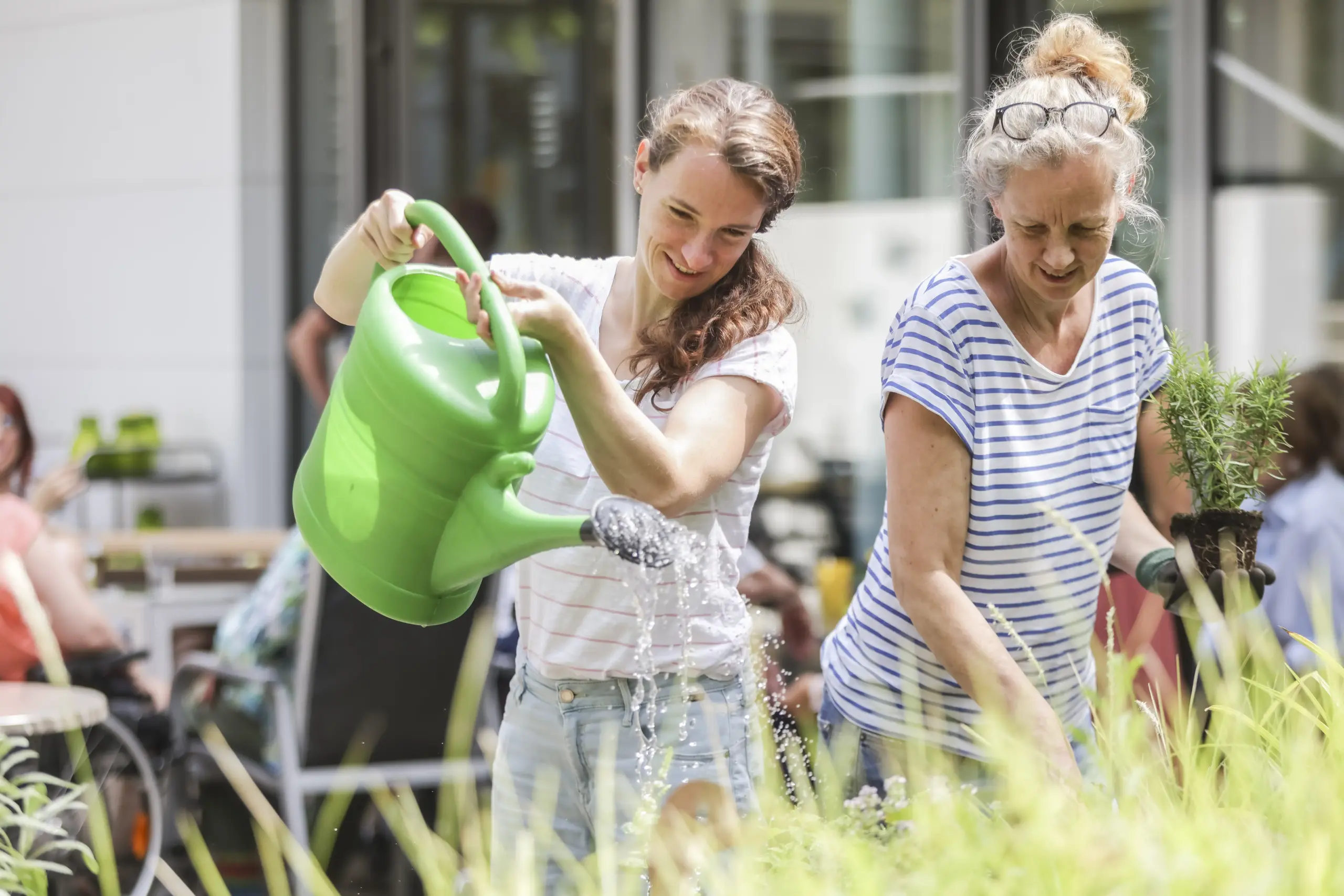 Zwei Frauen stehen nebeneinander bei der Gartenarbeit. Eine gießt mit einer grünen Gießkanne die Pflanzen. Die andere Frau hält in ihrer linken Hand einen Strauch, während sie mir ihrer rechten Hand ein kleines Loch dafür gräbt, um es einzupflanzen.