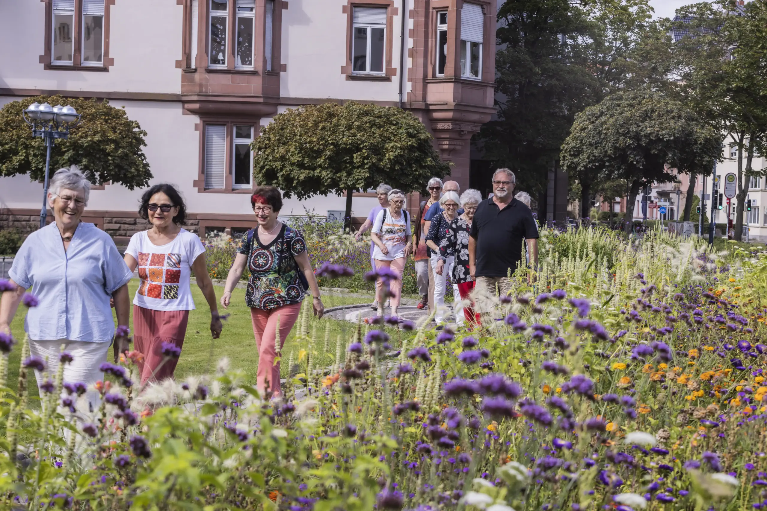 Quelle: BLV Eine Gruppe von etwa zehn Personen läuft hintereinander durch einen Park, vorbei an hohen Blumen. Im Hintergrund sind alte Häuser.