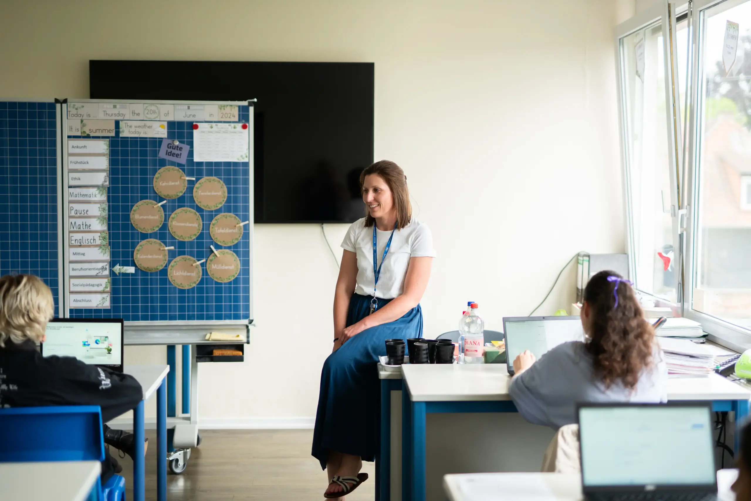Eine Lehrerein steht vor ihrer Klasse. Zwei Schülerinnen sitzen an den Tischen vor ihr und haben ihre Laptops geöffnet. Im Hintergrund steht eine Tafel.