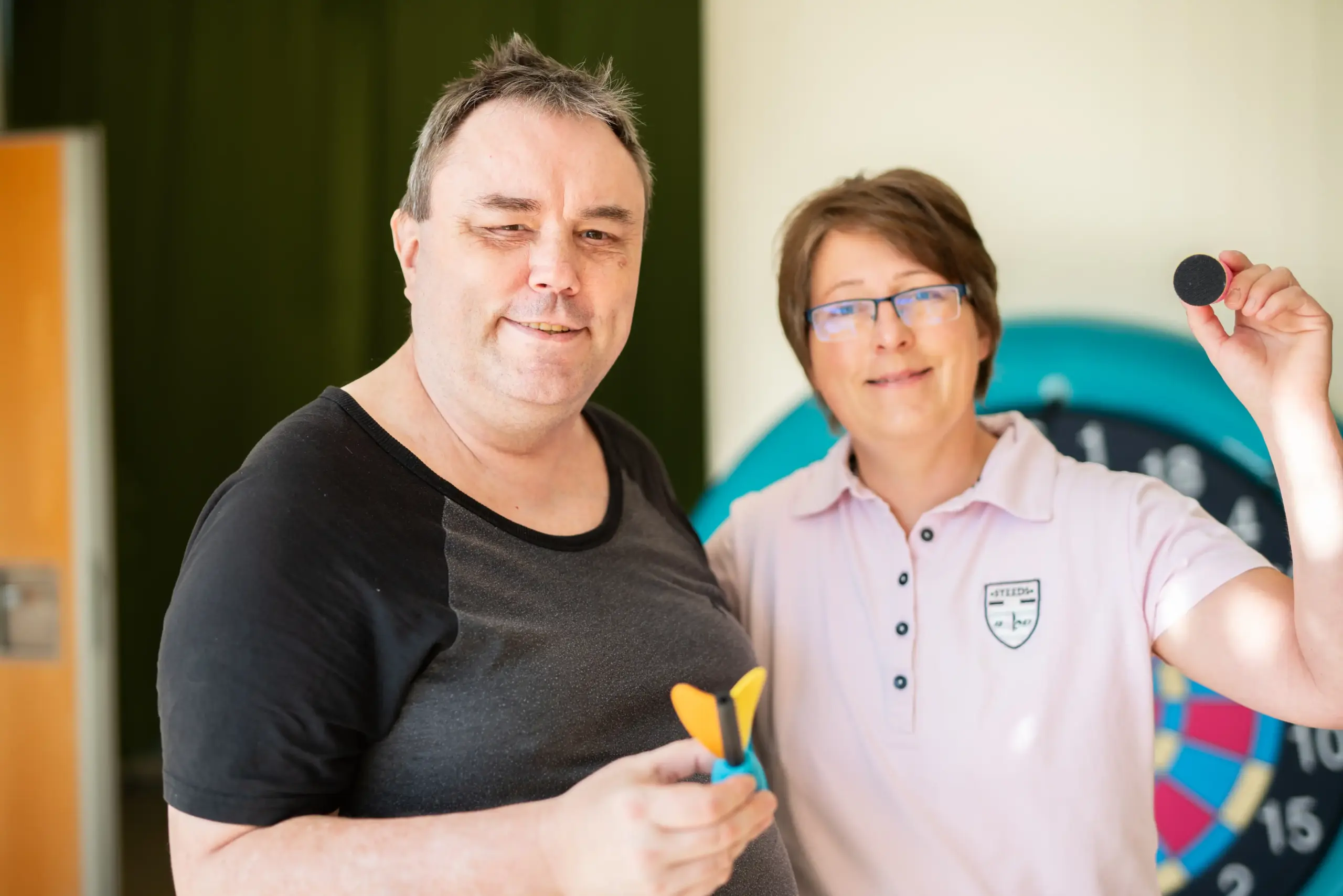 Ein Mann und eine Frau stehen nebeneinander. Sie präsentieren Dart-Pfeile. Im Hintergrund sieht man eine Dartscheibe.