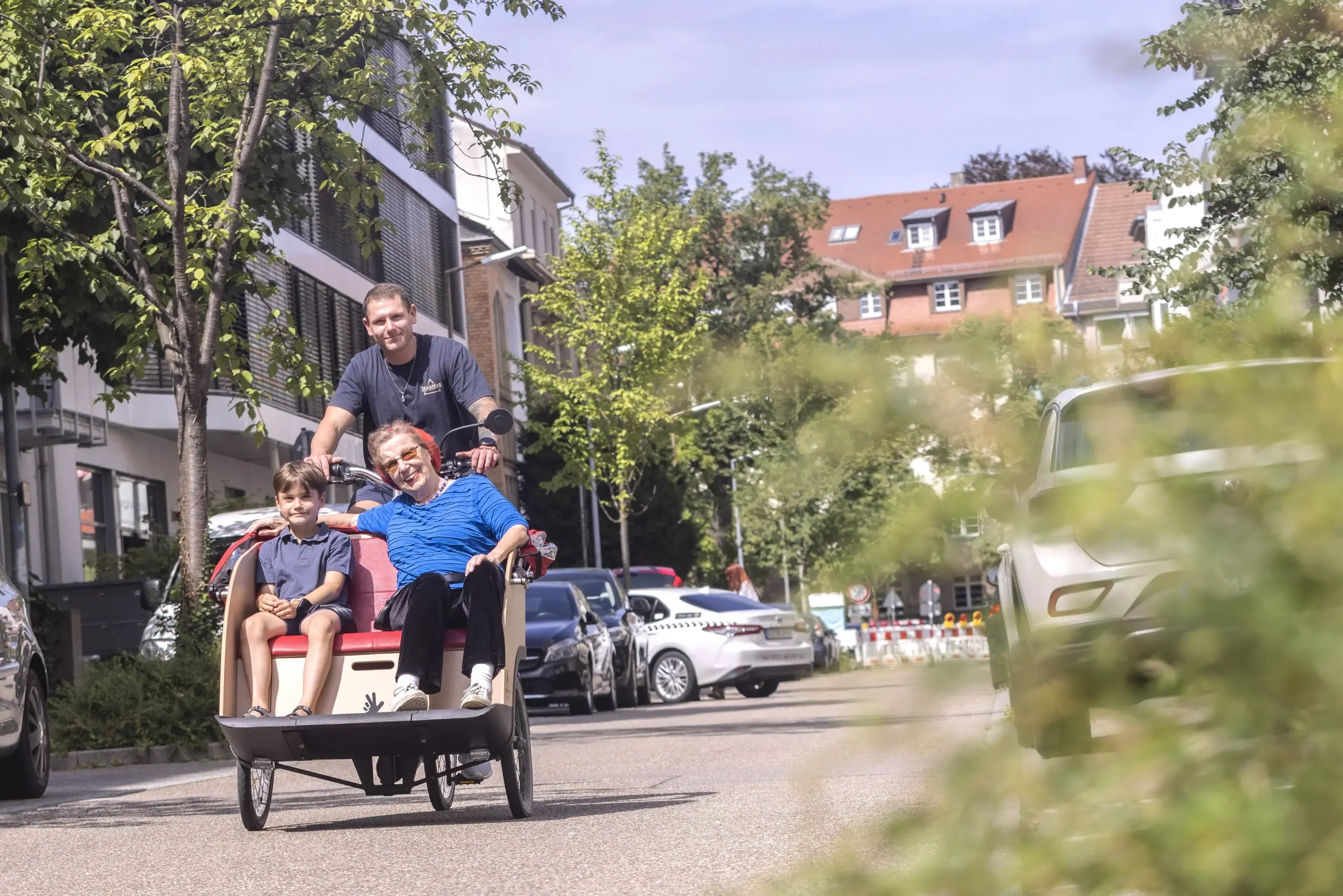 Ein Junge und eine ältere Frau sitzen in einer Rikscha. Ein Mann sitzt hinter dem Lenkrad und tritt in die Pedale.