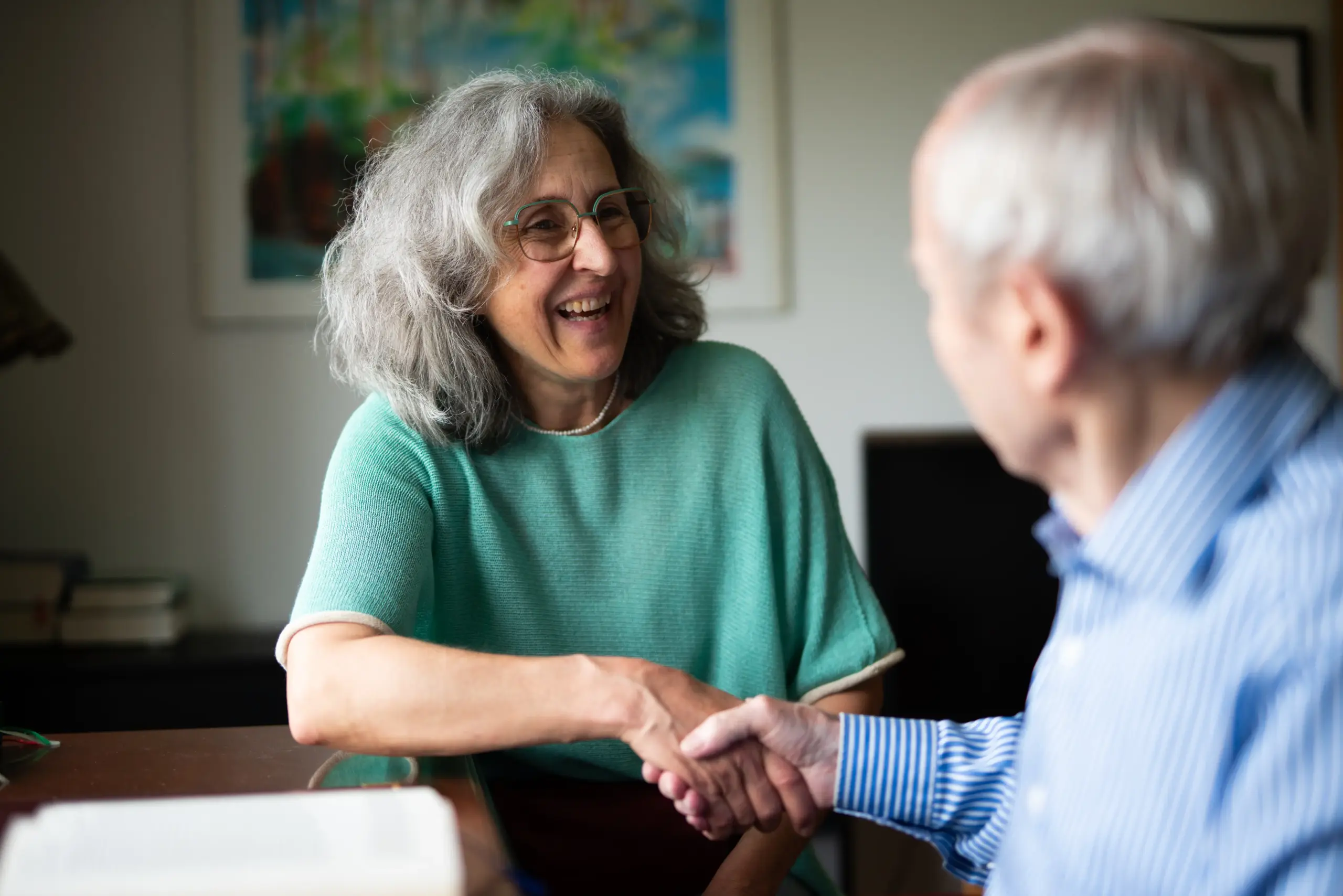 Zwei ältere Personen sitzen sich gegenüber und geben sich die Hand. Sie lachen.