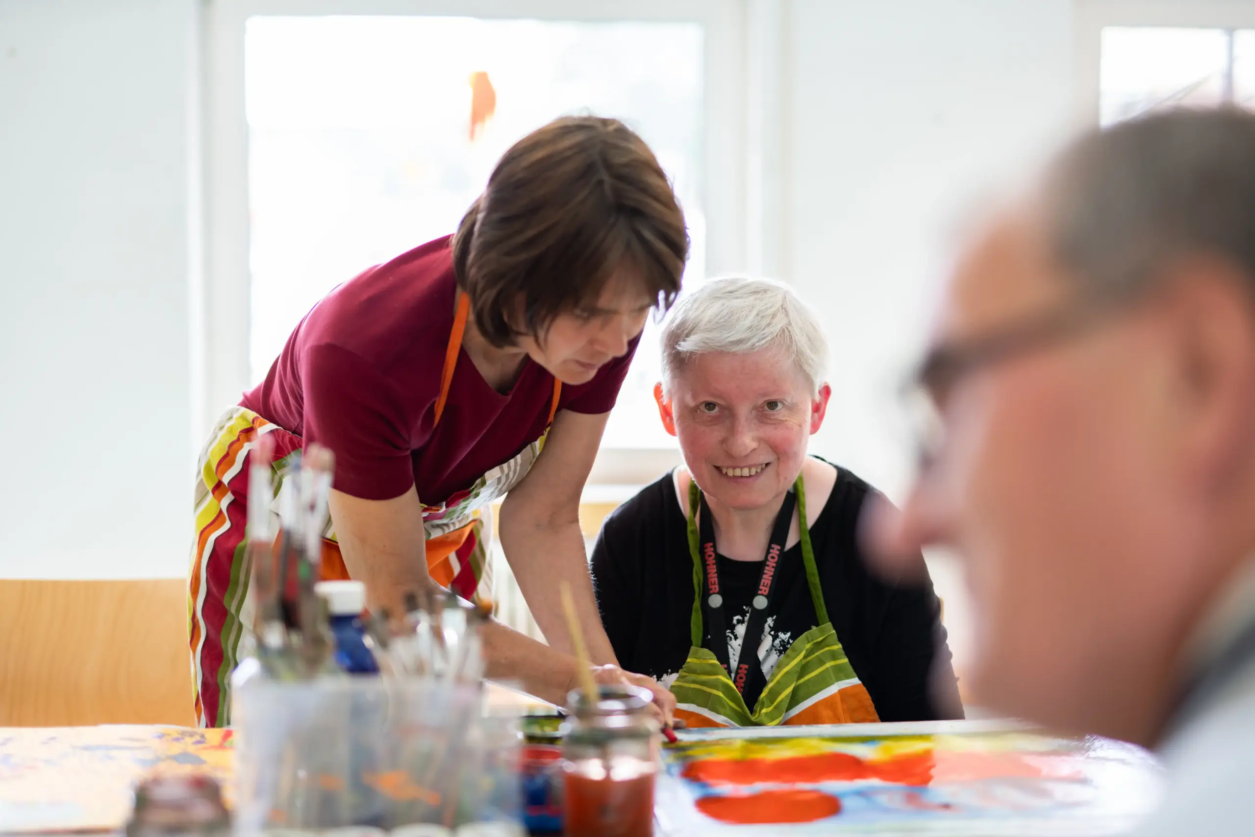 Quelle: BLV Eine Frau sitzt an einem Tisch und malt. Eine andere beugt sich über sie und hilft ihr dabei. Beide tragen bunte Schürzen.