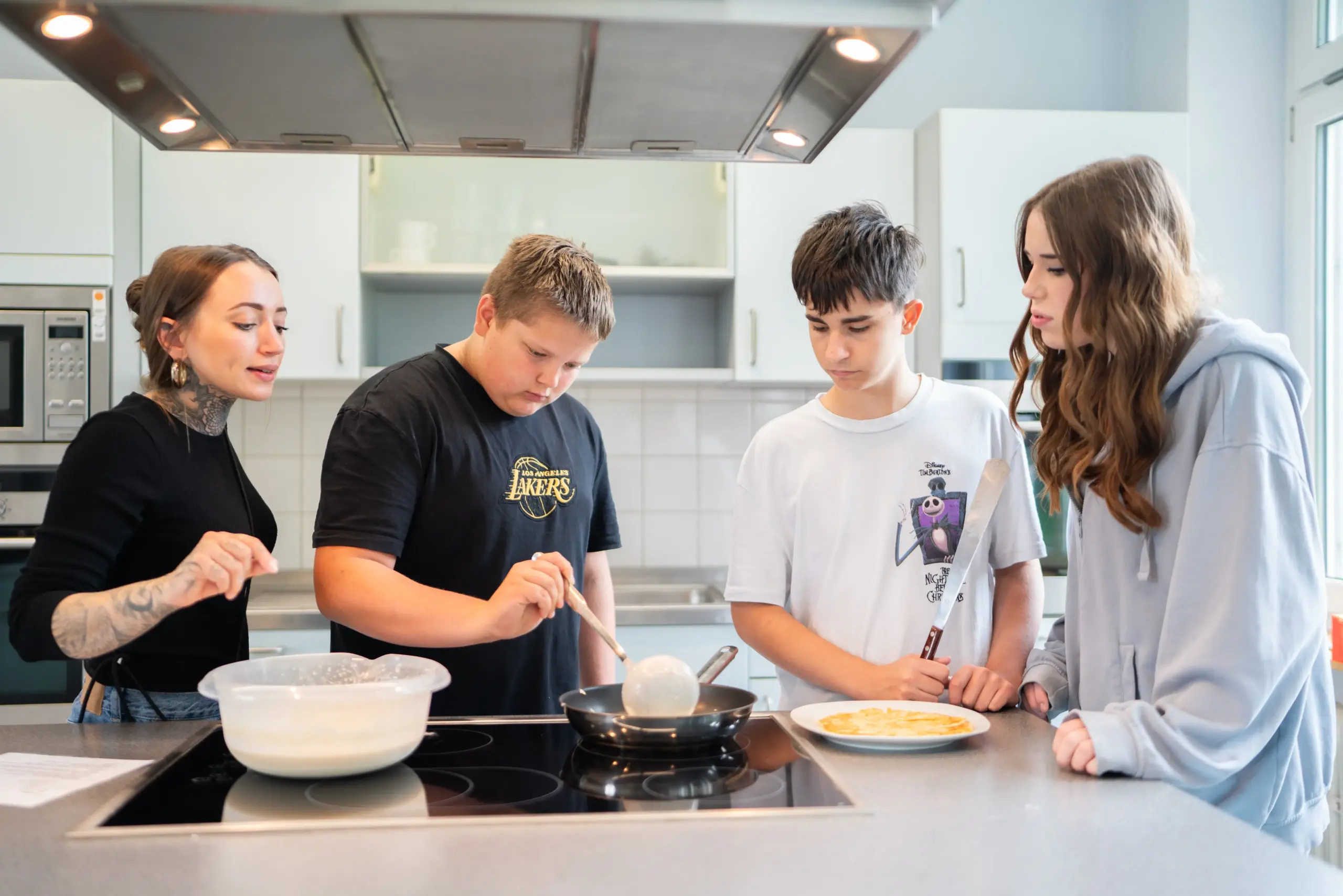Quelle: BLV Eine Frau und drei Jugendliche stehen in einer Großküche am Herd. Einer der Jungen schöpft mit einer Kelle Teig für einen Pfannenkuchen in die Pfanne. Die anderen schauen zu.