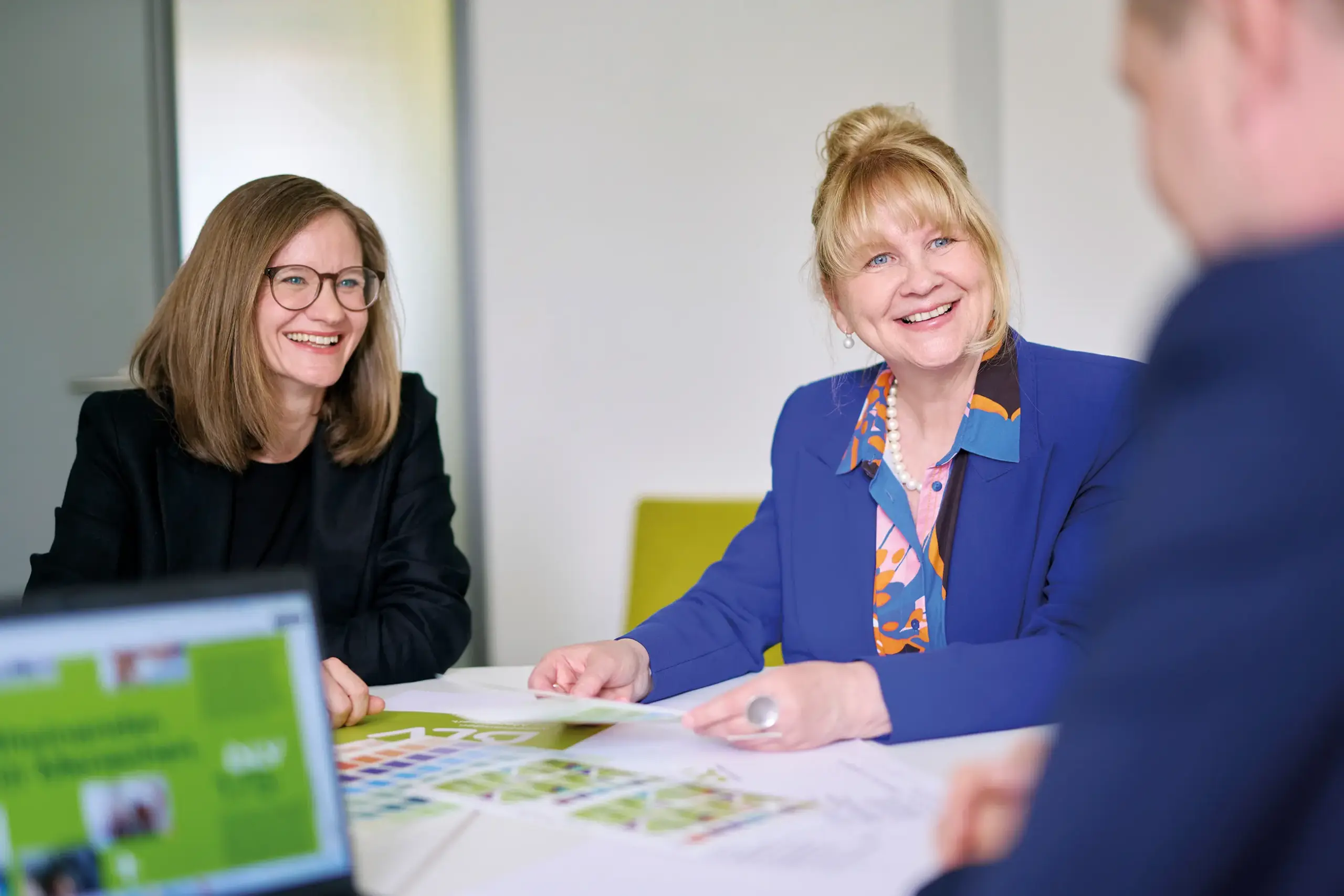 Drei Personen sitzen an einem Tisch. Die beiden Frauen lachen und schauen die dritte Person an.
