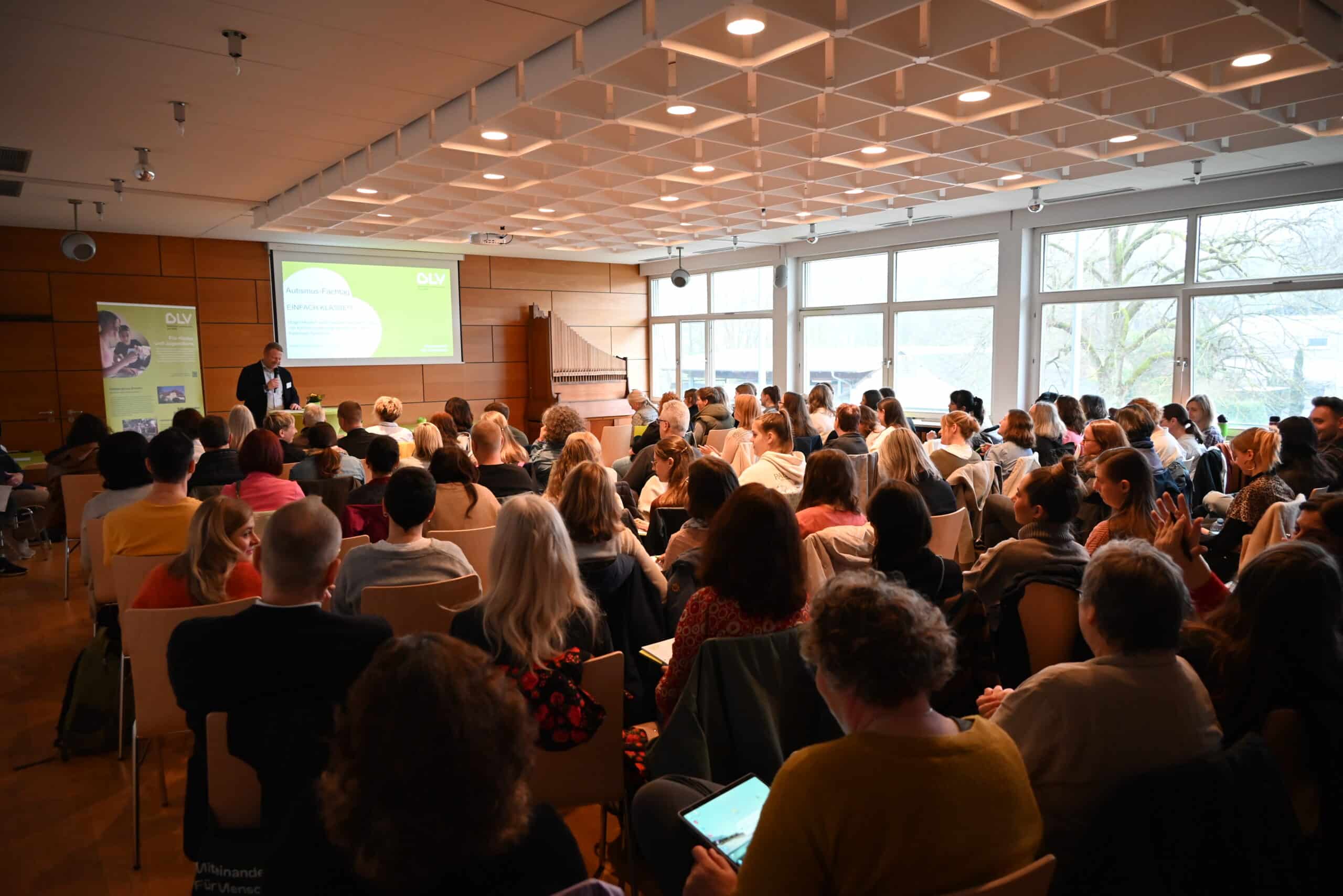 Christoph Röckinger hält anlässlich des "Fachtag Autismus" eine Rede vor großem Publikum im Hohberghaus. Im Hintergrund wird eine Präsentation auf eine Leinwand gestrahlt. Die Menschen im Publikum sitzen auf Holzstühlen und hören zu.