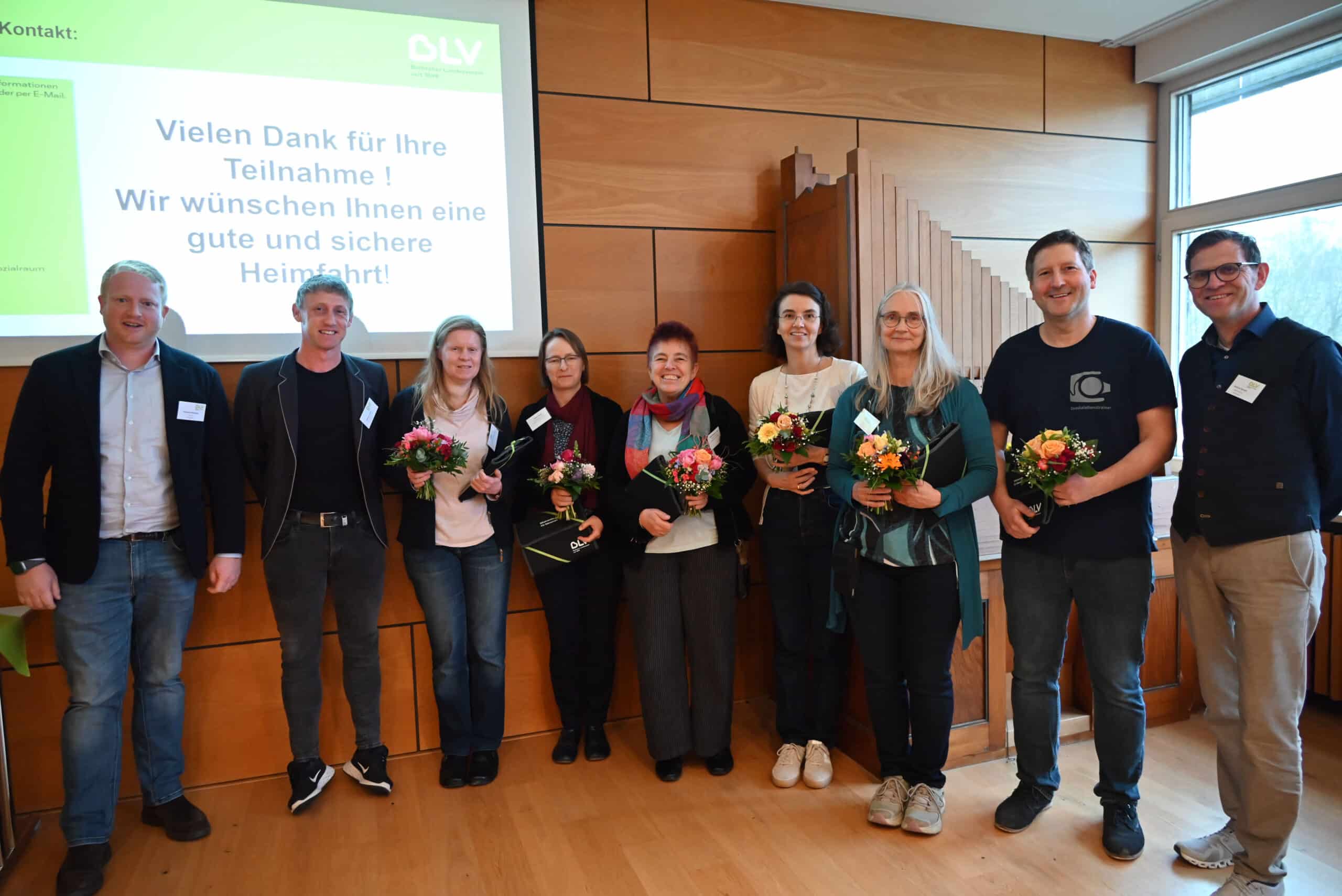Neun Personen stehen nebeneinander. Sechs von ihnen halten zum Dank für Ihre Vorträge beim 