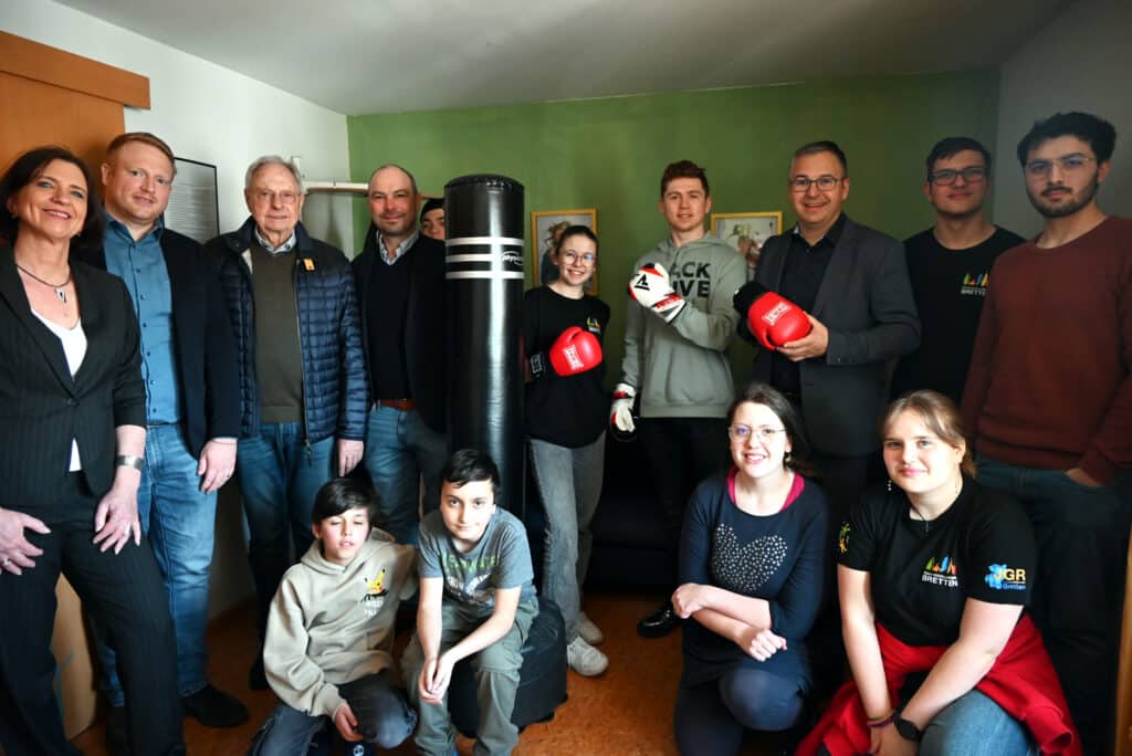 14 Personen rund um einen Boxsack. Vier junge Personen knien oder sitzen, der Rest steht dahinter. Drei von ihnen haben Boxhandschuhe an.