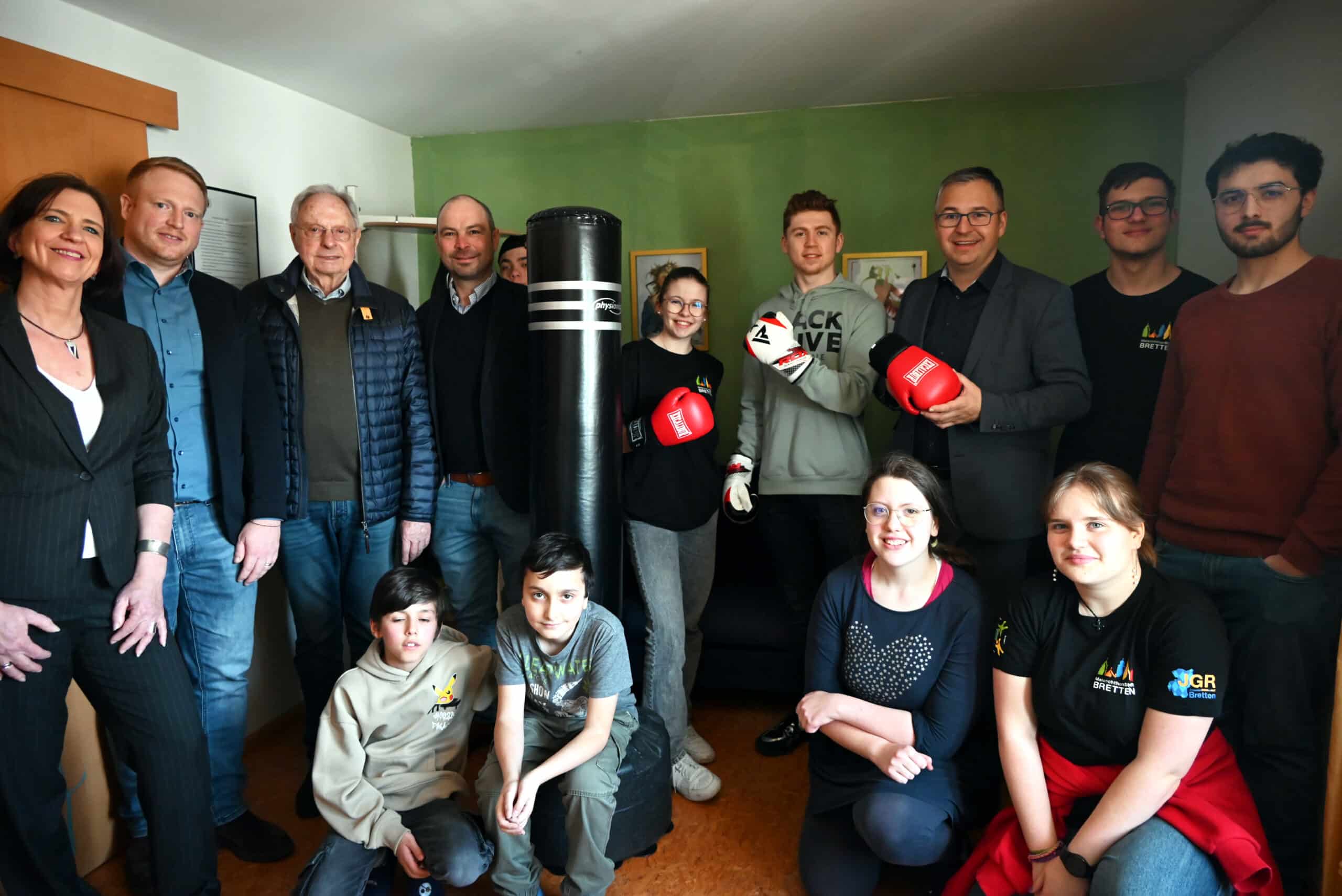 14 Personen rund um einen Boxsack. Vier junge Personen knien oder sitzen, der Rest steht dahinter. Drei von ihnen haben Boxhandschuhe an.