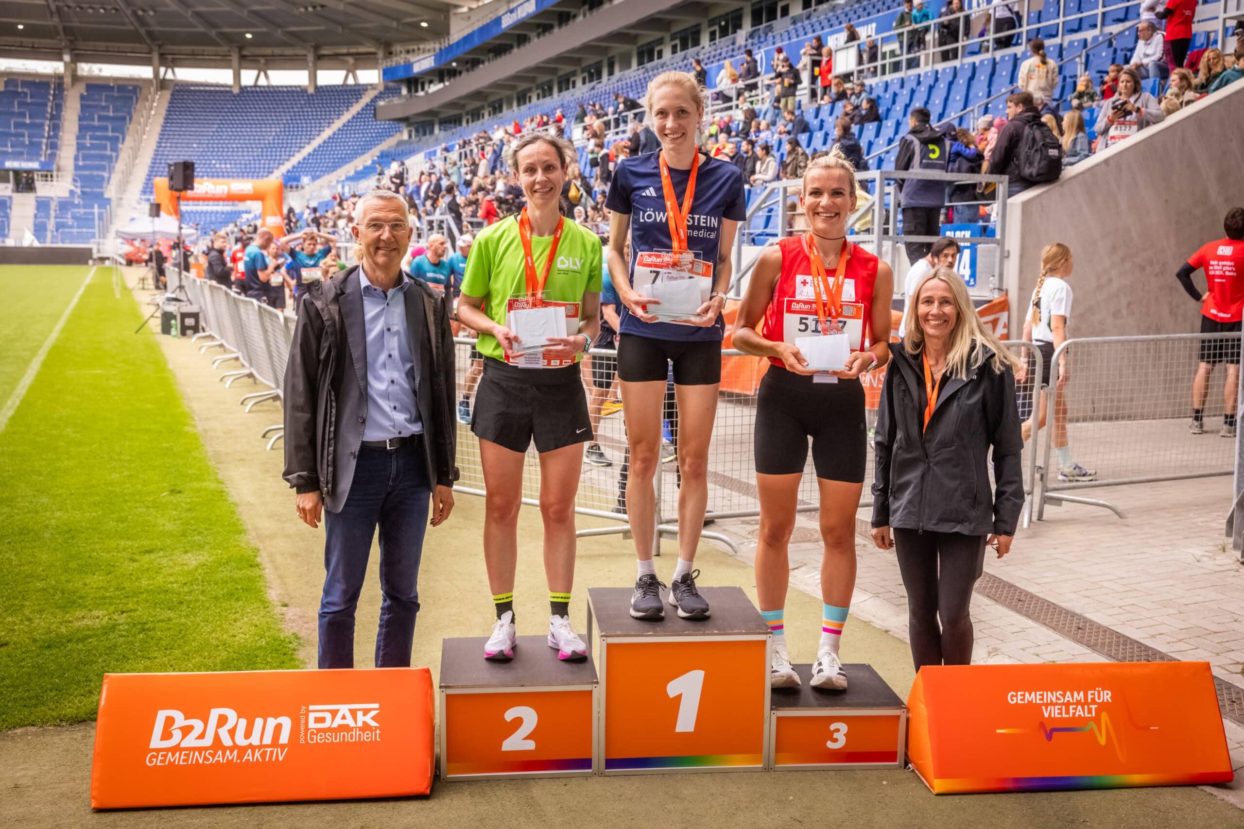 Quelle: Infront B2Run Auf einem Siegertreppchen stehen drei Frauen in Sportkleidung und mit Laufschuhen. Sie haben Medaillen um den Hals, halten ihre Preise in den Händen und lächeln. Links neben dem Podest ein Mann, der Bürgermeister Karlsruhes. Rechts neben dem Podest steht eine Frau. Im Hintergrund kann man die Tribüne eines Stadions erkennen. Hinter einer Absperrung kommen weitere Läuferinnen und Läufer ins Ziel.