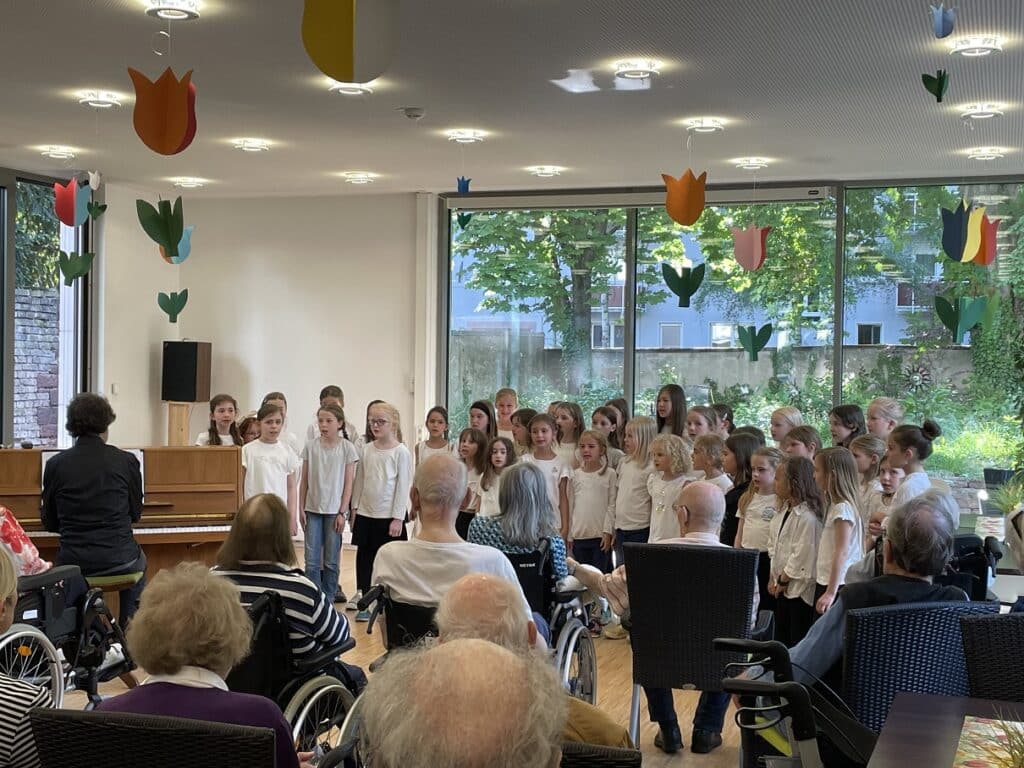 Ein Chor aus rund 4 jungen Mädchen singen vor einem Publikum. Sie tragen alle weiße T-Shirts. Ein Mann mit dunklem Hemd begleitet sie auf dem Klavier. Im Publikum sitzen vor allem ältere Personen, einige davon auch im Rollstuhl.