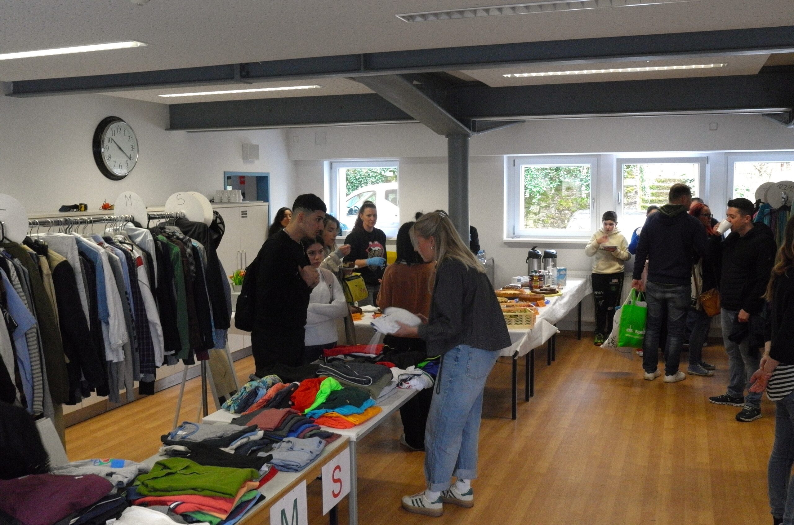 Quelle: BLV/Bischoff Drei Personen prüfen Kleidungsstücke an einem Tisch, auf dem noch viele weitere Kleidungsstücke ausgebreitet sind. Im Hintergrund steht ein Tisch mit deiversen Kuchen sowie Kaffeekannen und Milchpackungen. Mehrere Menschen essen Kuchen oder trinken aus Tassen.