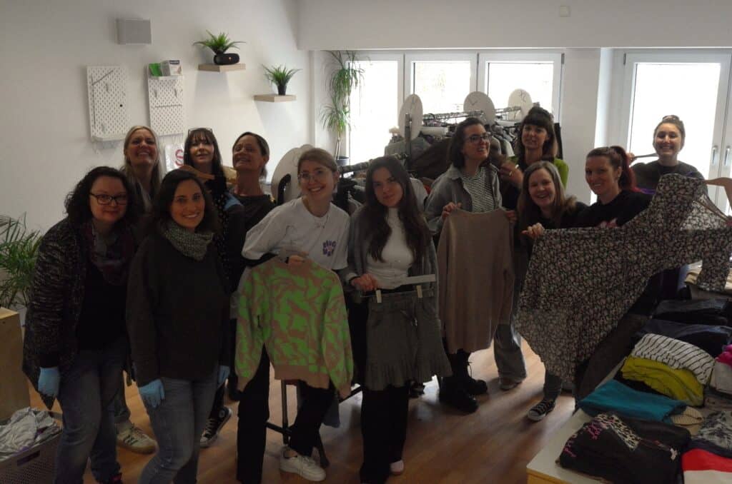 12 Frauen auf dem Kleiderbasar der Schule Enzberg. Einige von ihnen präsentieren unterschiedliche Kleidungsstücke. Auf einem Tisch liegen diverse Pullover. Im Hintergrund hängen an Kleiderstangen weitere Kleidungsstücke.