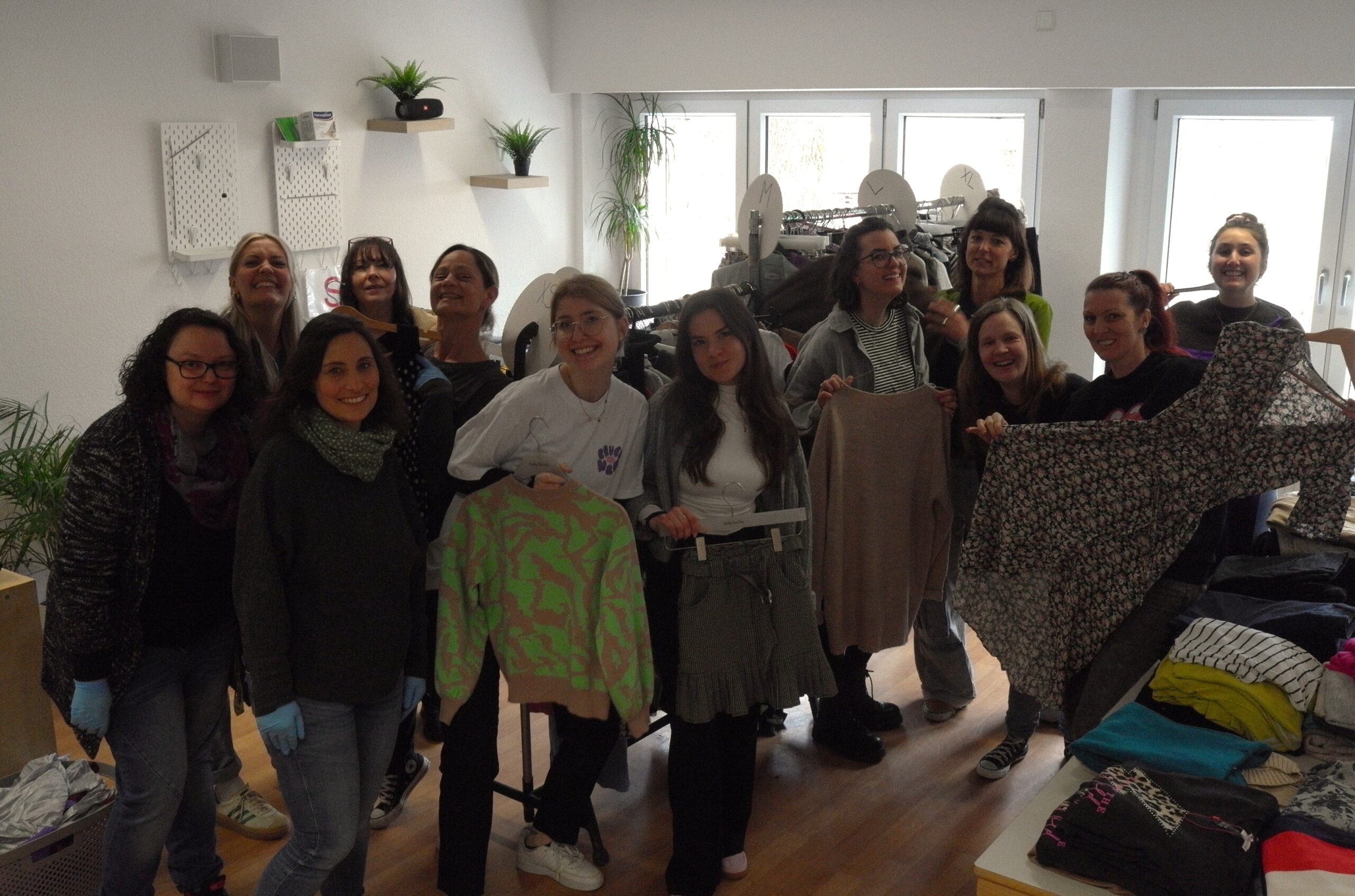 12 Frauen auf dem Kleiderbasar der Schule Enzberg. Einige von ihnen präsentieren unterschiedliche Kleidungsstücke. Auf einem Tisch liegen diverse Pullover. Im Hintergrund hängen an Kleiderstangen weitere Kleidungsstücke.