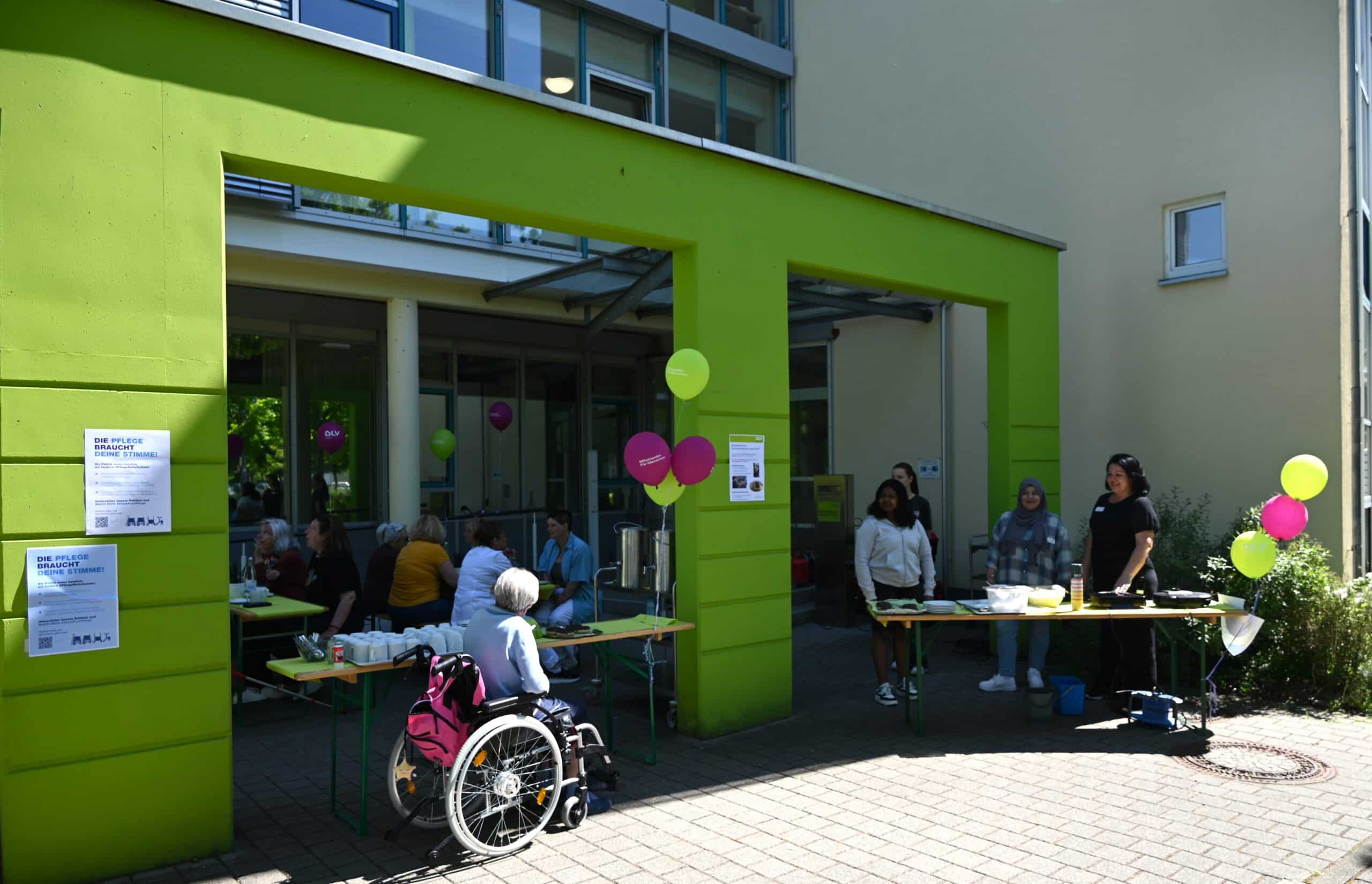 Vor dem Eingang des Altenhilfezentrums Karlsruhe-Nordost bieten drei Mitarbeiterinnen der Einrichtung an zwei Tischen Kaffee und Waffeln an. Eine Frau im Rollstuhl betrachtet das Angebot. Im Hintergrund sitzen mehrere Menschen an Biertischen und unterhalten sich.