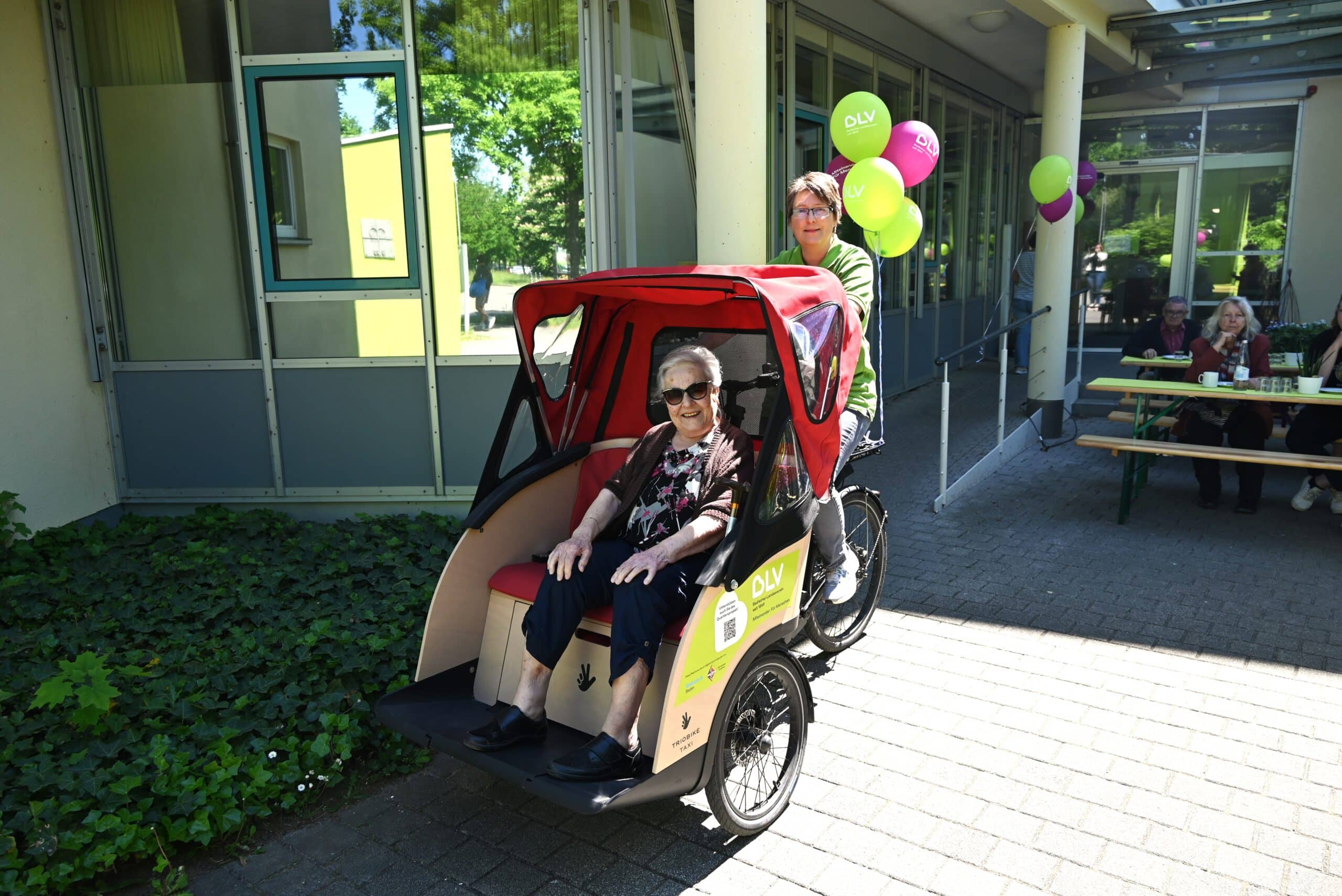 Eine ältere Frau sitzt in einer Rikscha. Sie trägt eine Sonnenbrille und lacht. Die Rikscha wird von einer jüngeren Frau im BLV-T-Shirt gefahren. Im Hintergrund sieht man einige grüne und violette Luftballons sowie mehrere Biertischgarnituren an denen ein paar Personen sitzen.