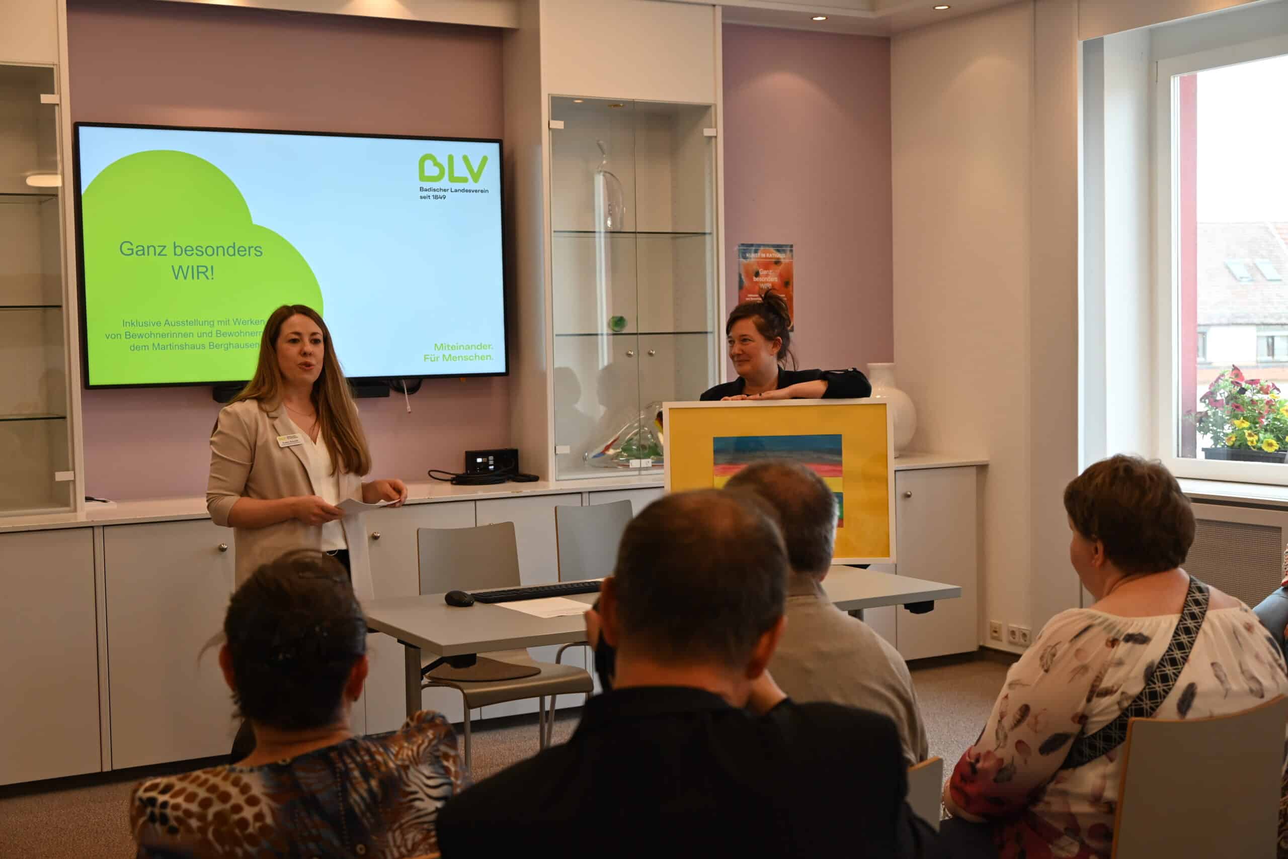 Evelyn Schmidt, Einrichtungsleitung Martinshaus, hält eine Rede im Pfinztaler Rathaus. Neben ihr steht Nicola Bodner, Bürgermeisterin der Gemeinde Pfinztal. Hinter den beiden sieht man auf einem großen Bildschirm eine Präsentation mit dem Thema der Veranstaltung.