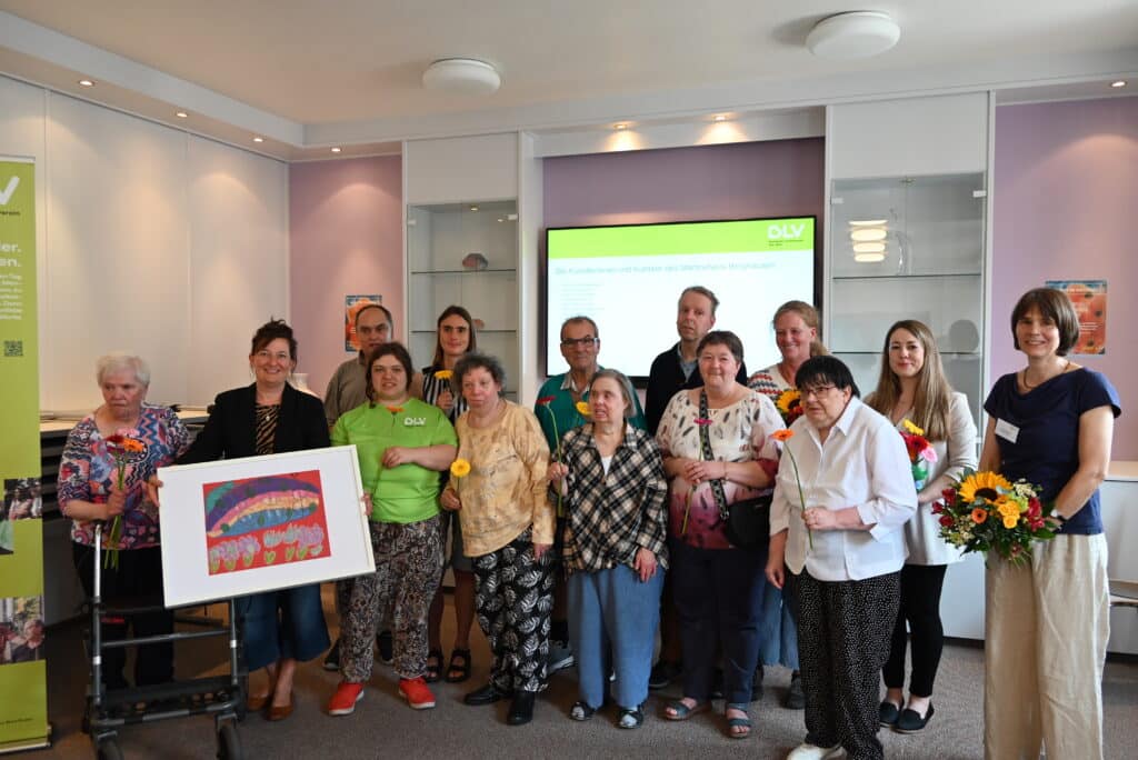 Gruppenbild der Künstlerinnen und Künstler des Martinshauses Berghausen mit Bürgermeisterin Nicola Bodner (2.v.l.) sowie Evelyn Schmidt, der Einrichtungsleitung und weiteren Mitarbeitenden des Martinshauses. Alle Personen haben eine Blume in der Hand.