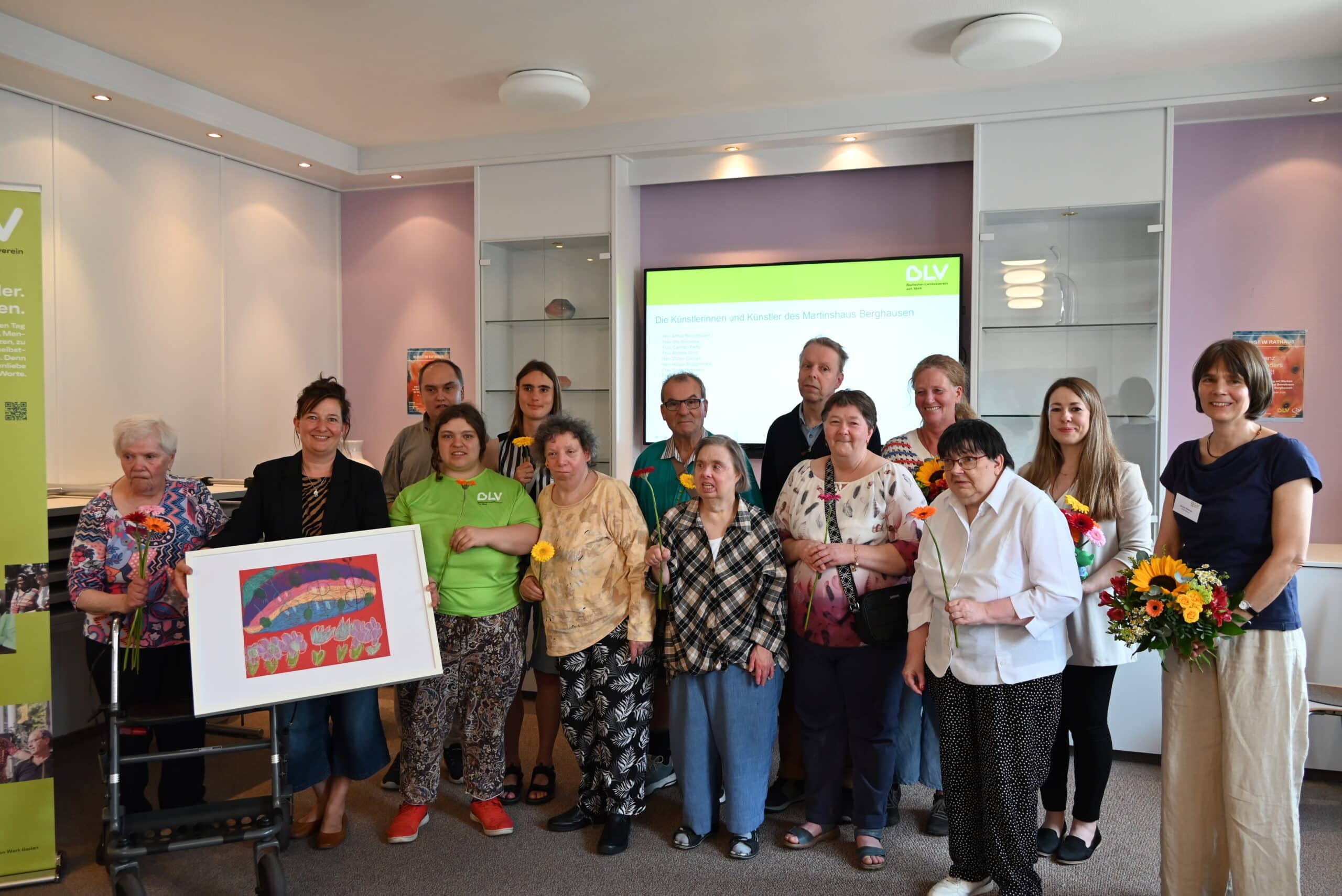 Gruppenbild der Künstlerinnen und Künstler des Martinshauses Berghausen mit Bürgermeisterin Nicola Bodner (2.v.l.) sowie Evelyn Schmidt, der Einrichtungsleitung und weiteren Mitarbeitenden des Martinshauses. Alle Personen haben eine Blume in der Hand.