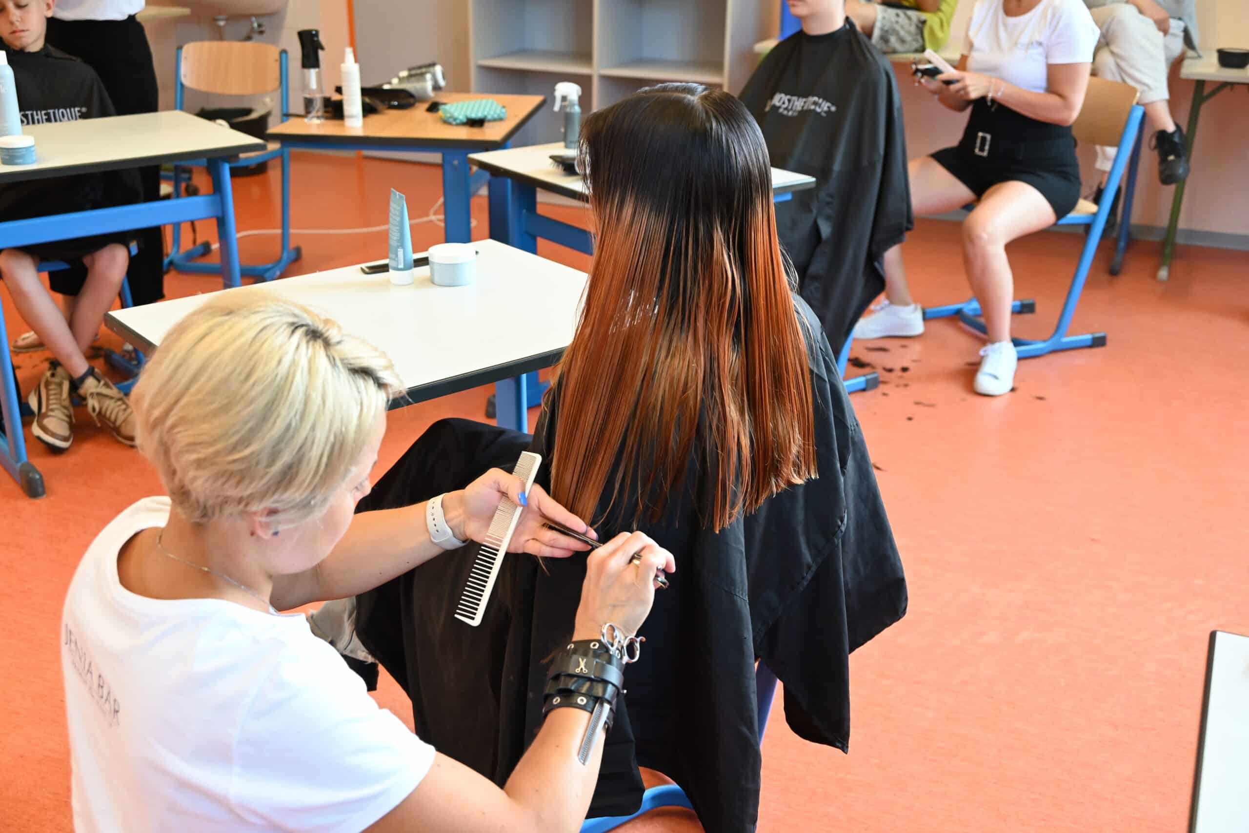 Ein Mädchen mit schwarzen Haaren und rotbraunen Spitzen sitzt in einem Klassenzimmer auf einem Stuhl. Ihr werden von einer Frau mit blonden Haaren und weißem T-Shirt die Spitzen geschnitten. Die Frau hält eine Schere und einen Kamm in ihren Händen. Im Hintergrund werden weiteren Kindern die Haare geschnitten.